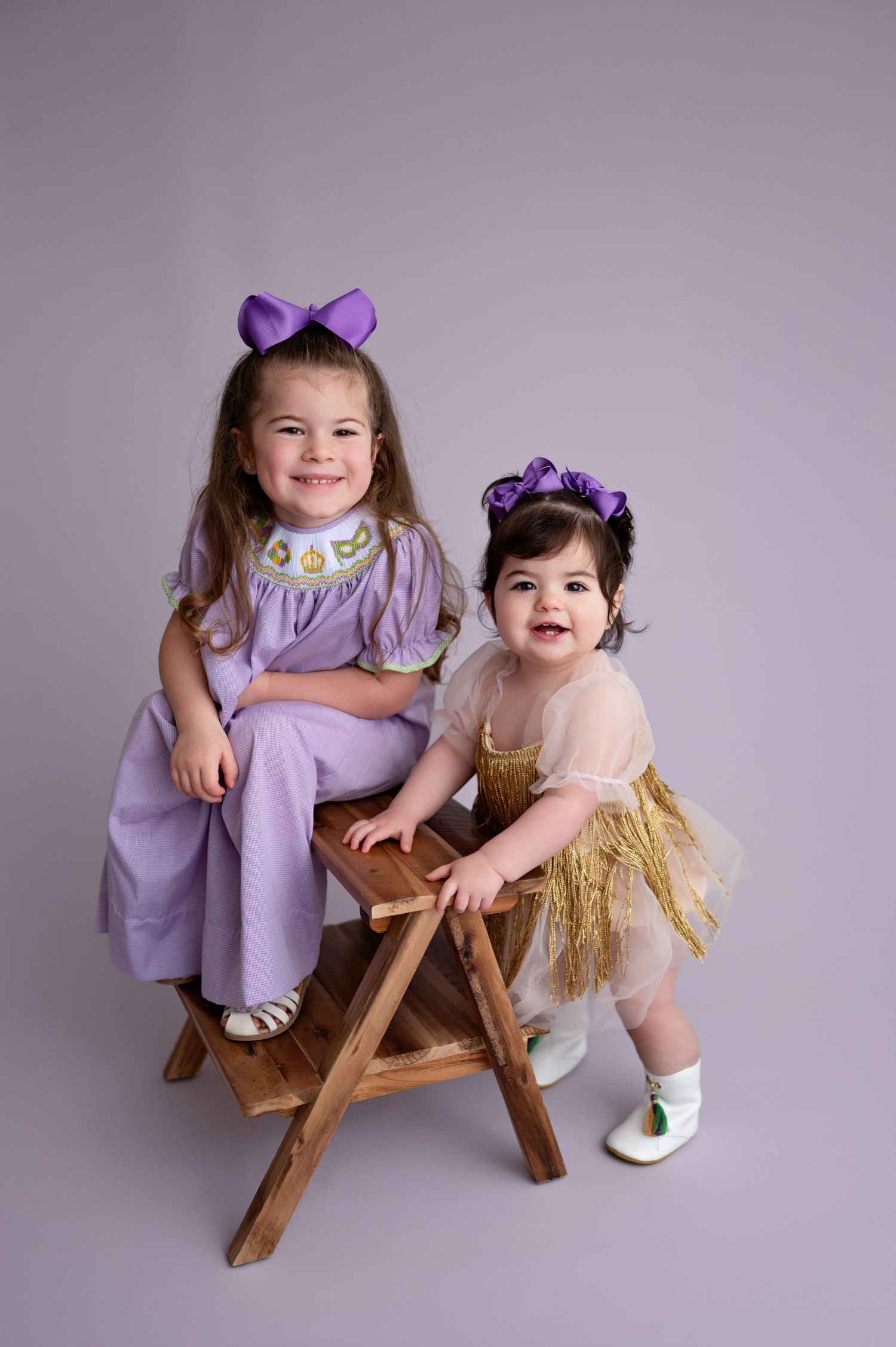 Two children in purple and pink dresses sit and play together on a wooden bench in a studio setting.