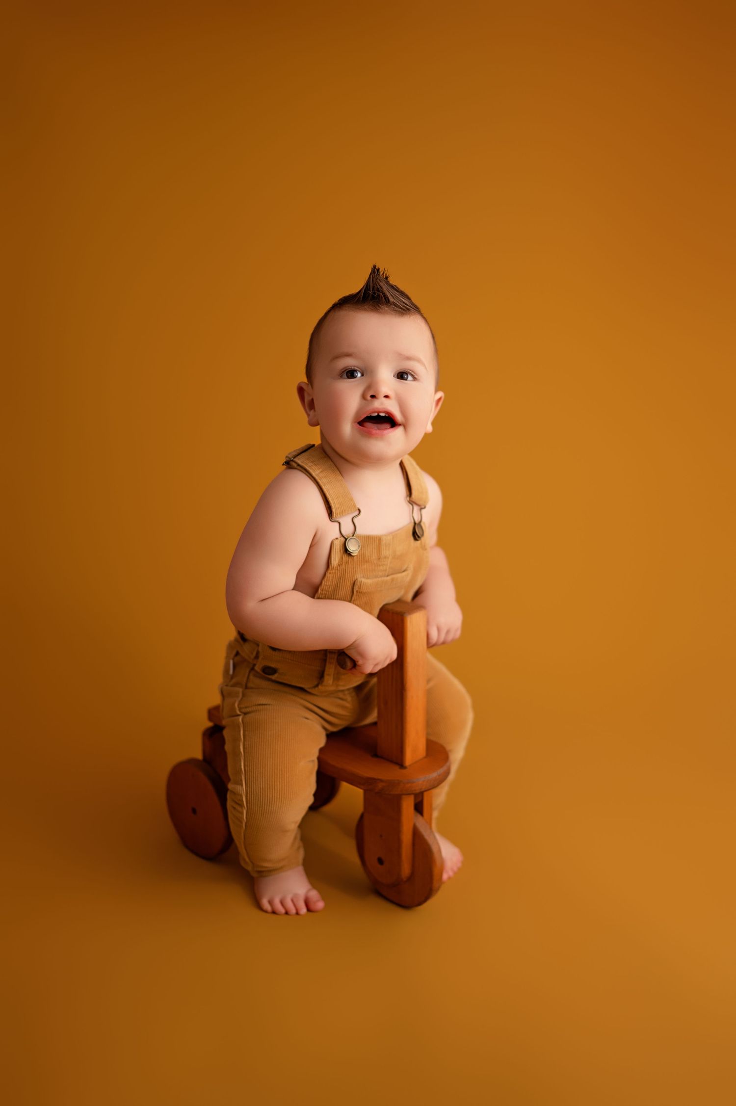 Baby wearing brown overalls sits on wooden toy bike against orange background.