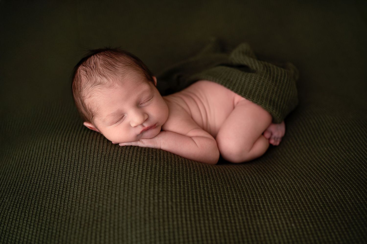 A sleeping newborn baby rests peacefully on a green textured blanket in dim lighting.