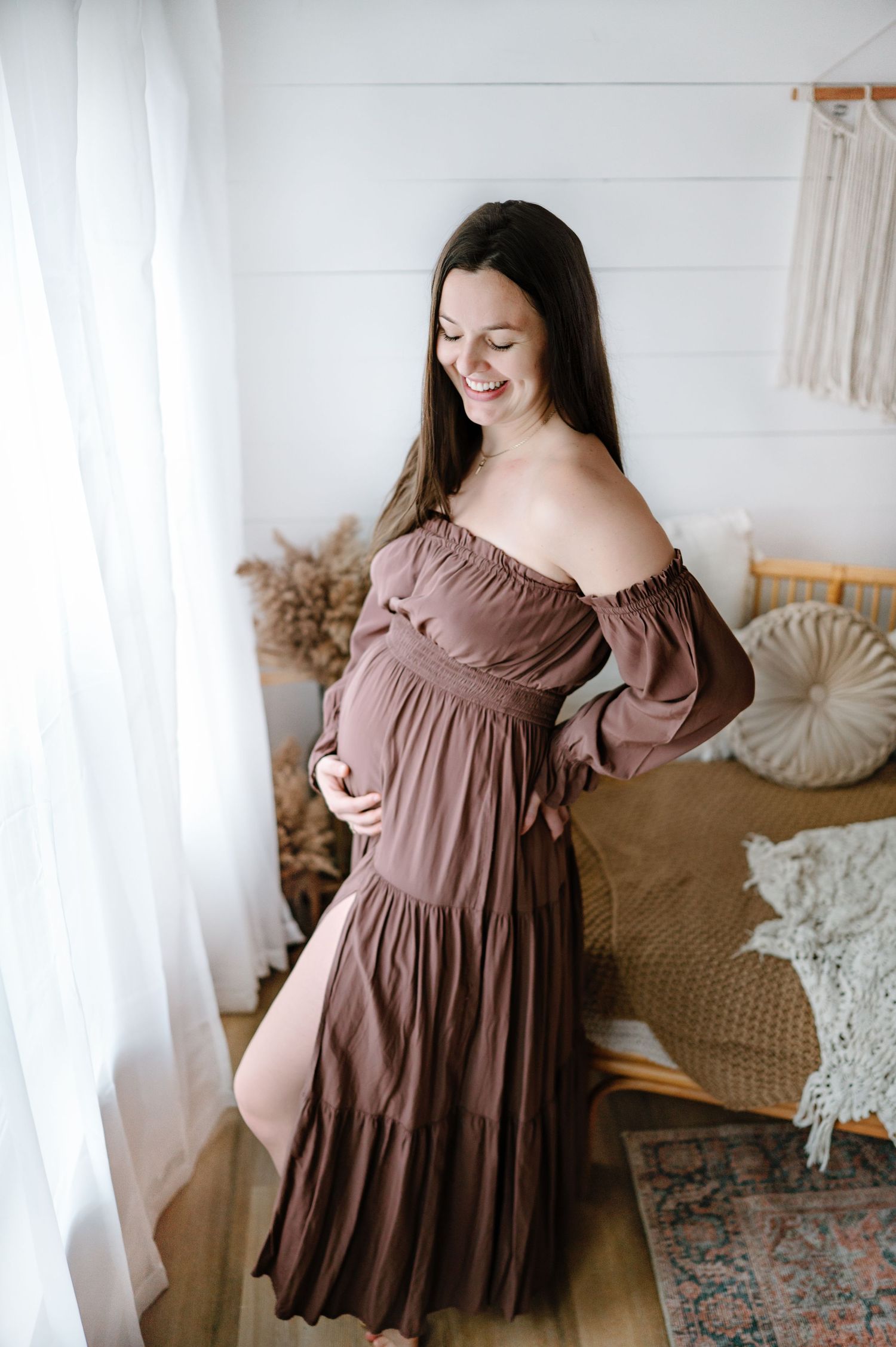 Pregnant woman in maxi dress smiles while posing in a bright, boho-styled room with white walls and natural decor.