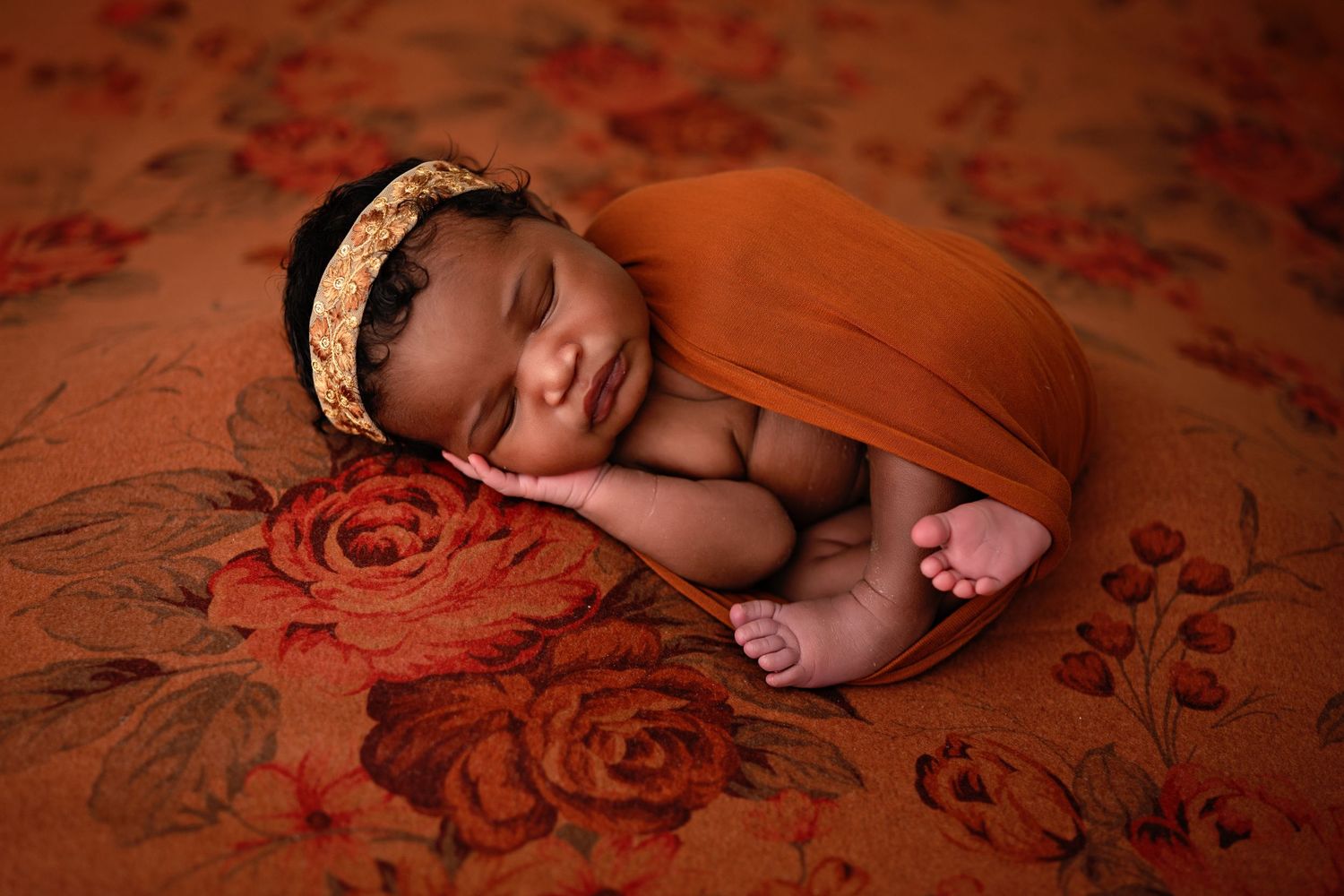 Sleeping newborn baby wearing floral headband lying on red floral backdrop during professional photo session.