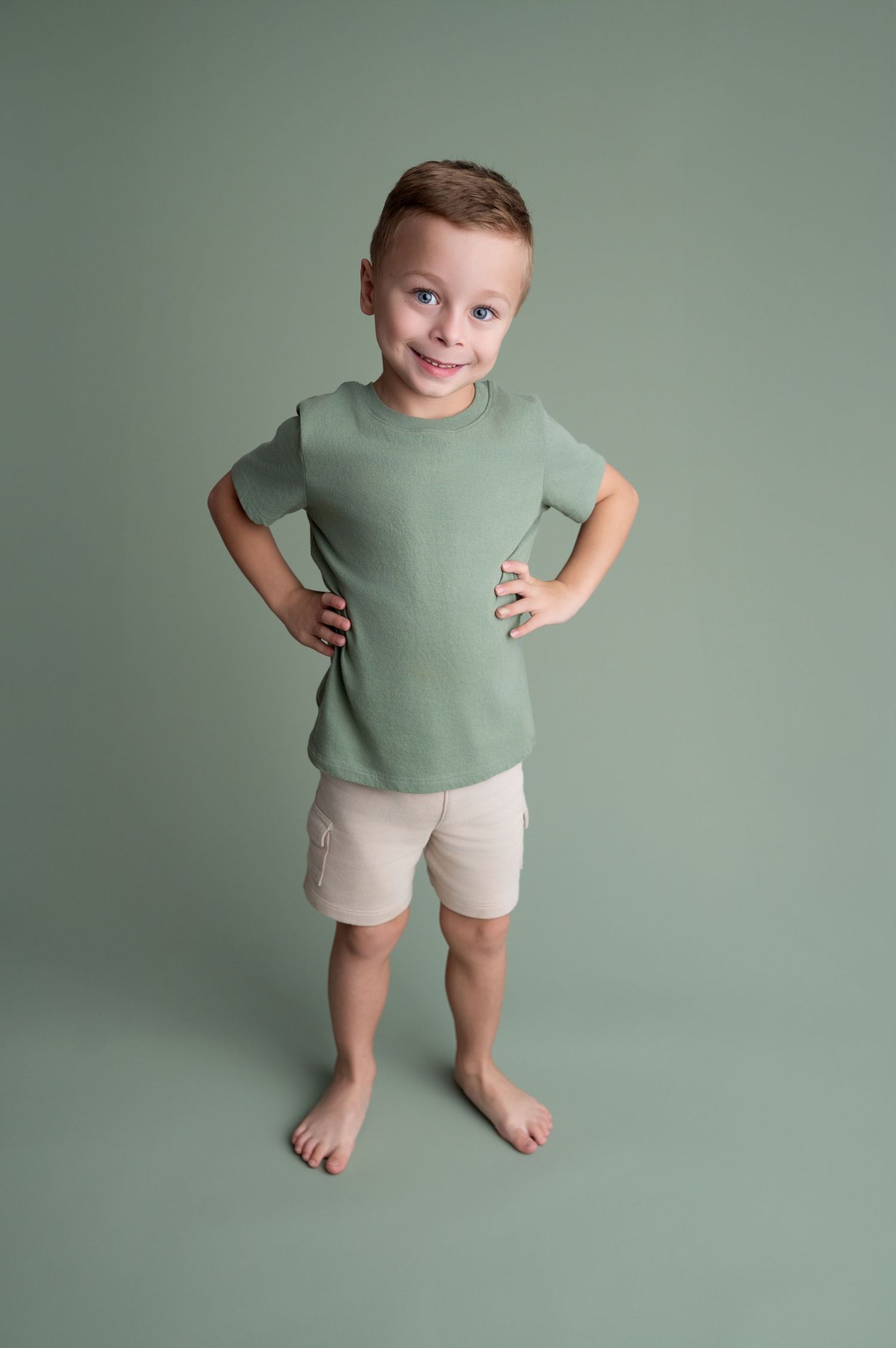 Small child wearing sage green t-shirt and beige shorts stands against mint green backdrop in playful poses.