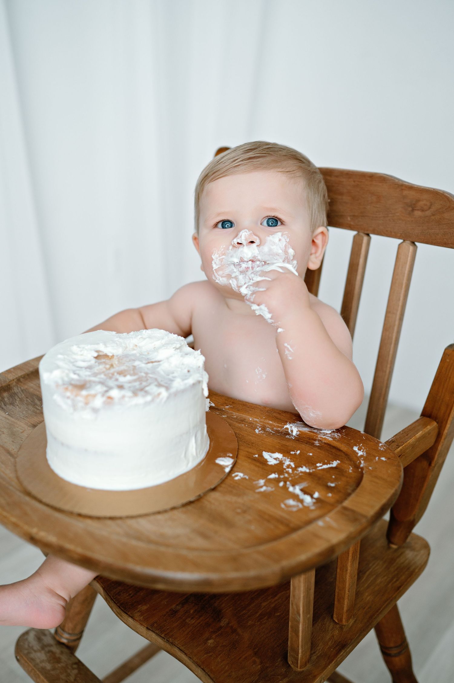 Baby eating birthday cake gets messy with white frosting in wooden high chair during first birthday celebration.