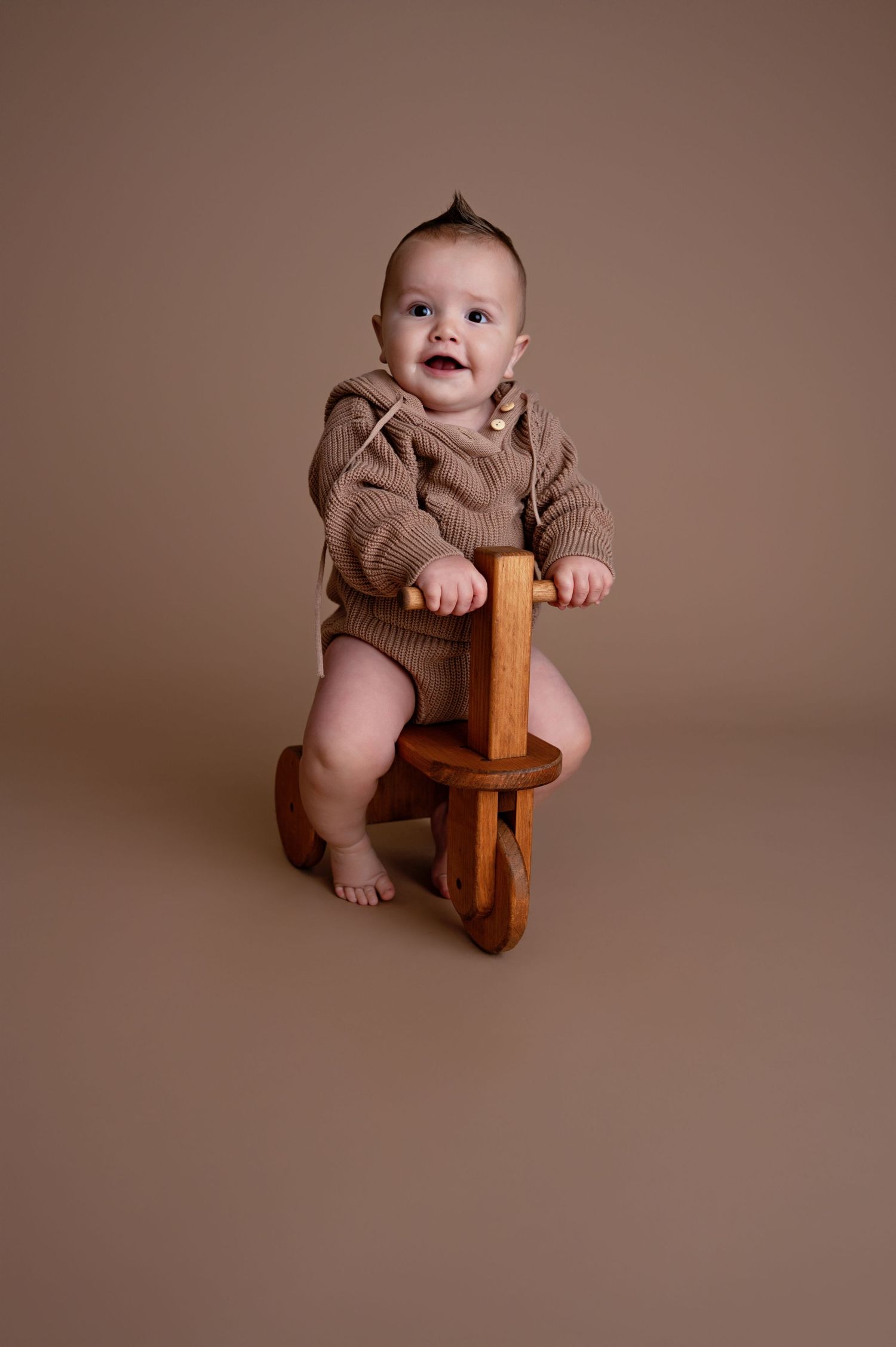 A adorable baby wearing brown knitwear sits on a wooden stool against a warm brown studio backdrop.