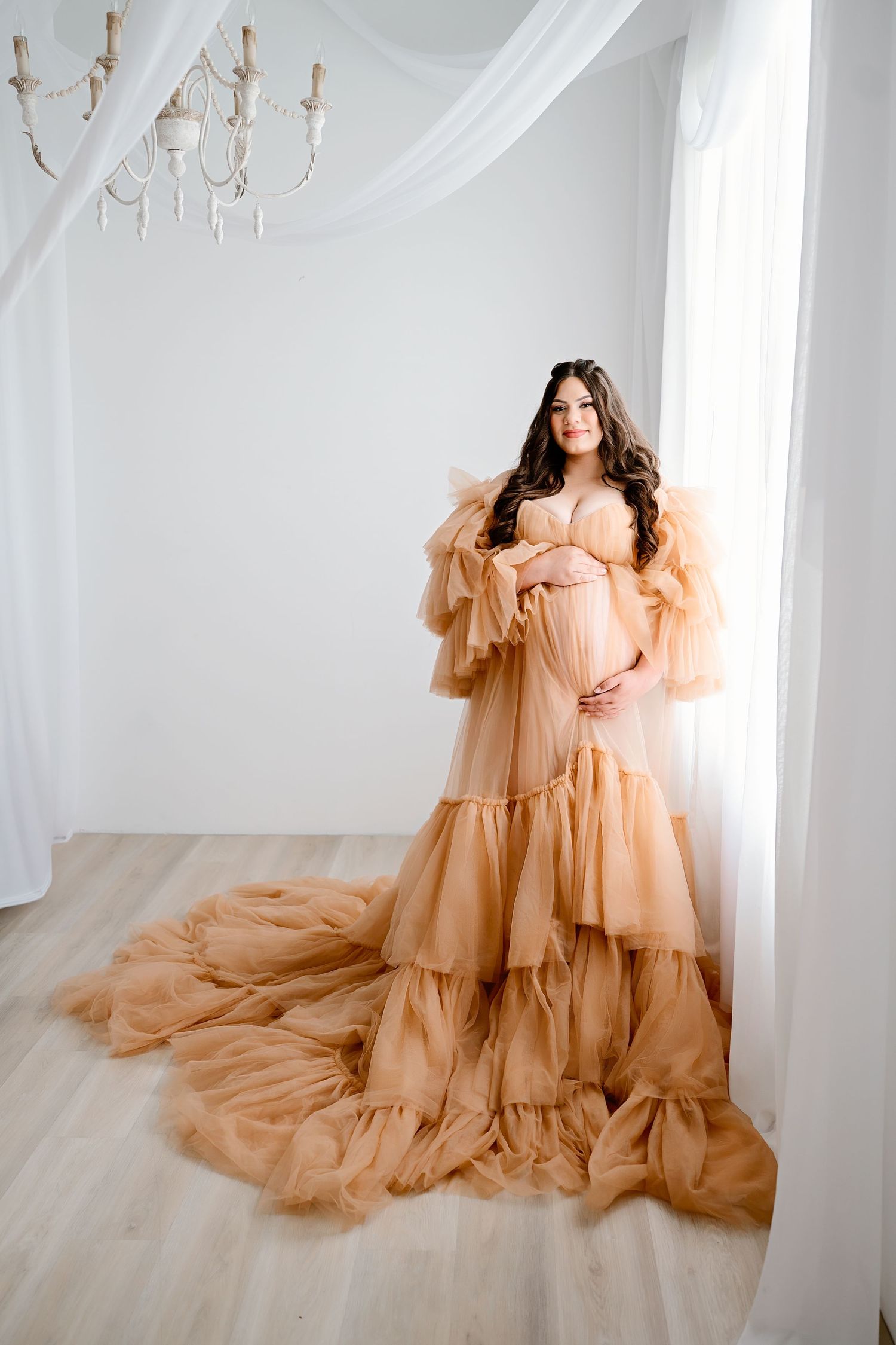 Elegant maternity photo featuring flowing champagne tulle dress in white room with chandelier.
