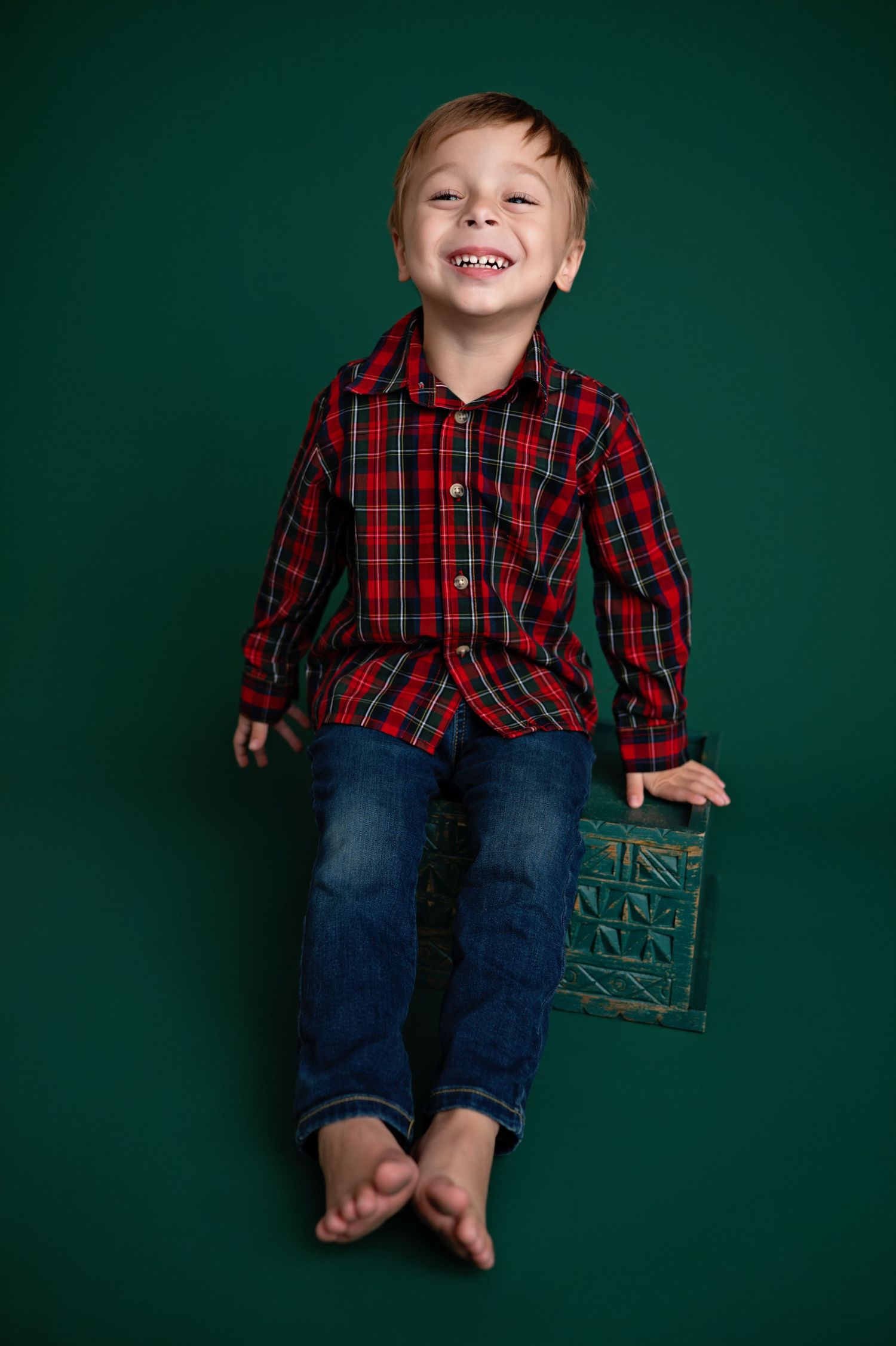 Child wearing red plaid shirt and jeans sits against dark green wall in casual holiday portrait series.