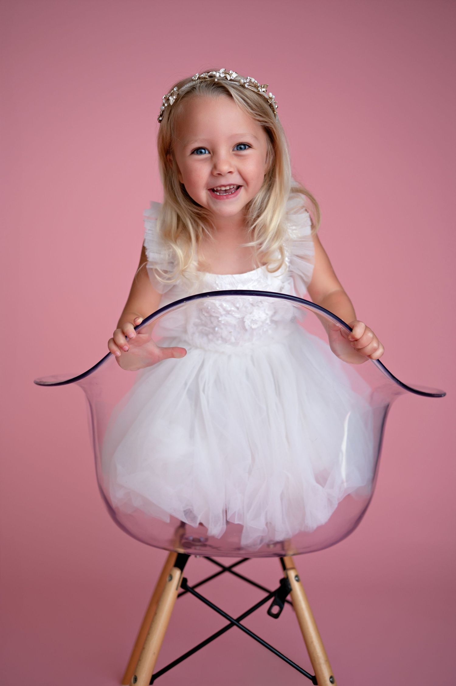 Child in white tulle dress poses cheerfully on modern Eames-style chair against pink backdrop in sequence of photos.