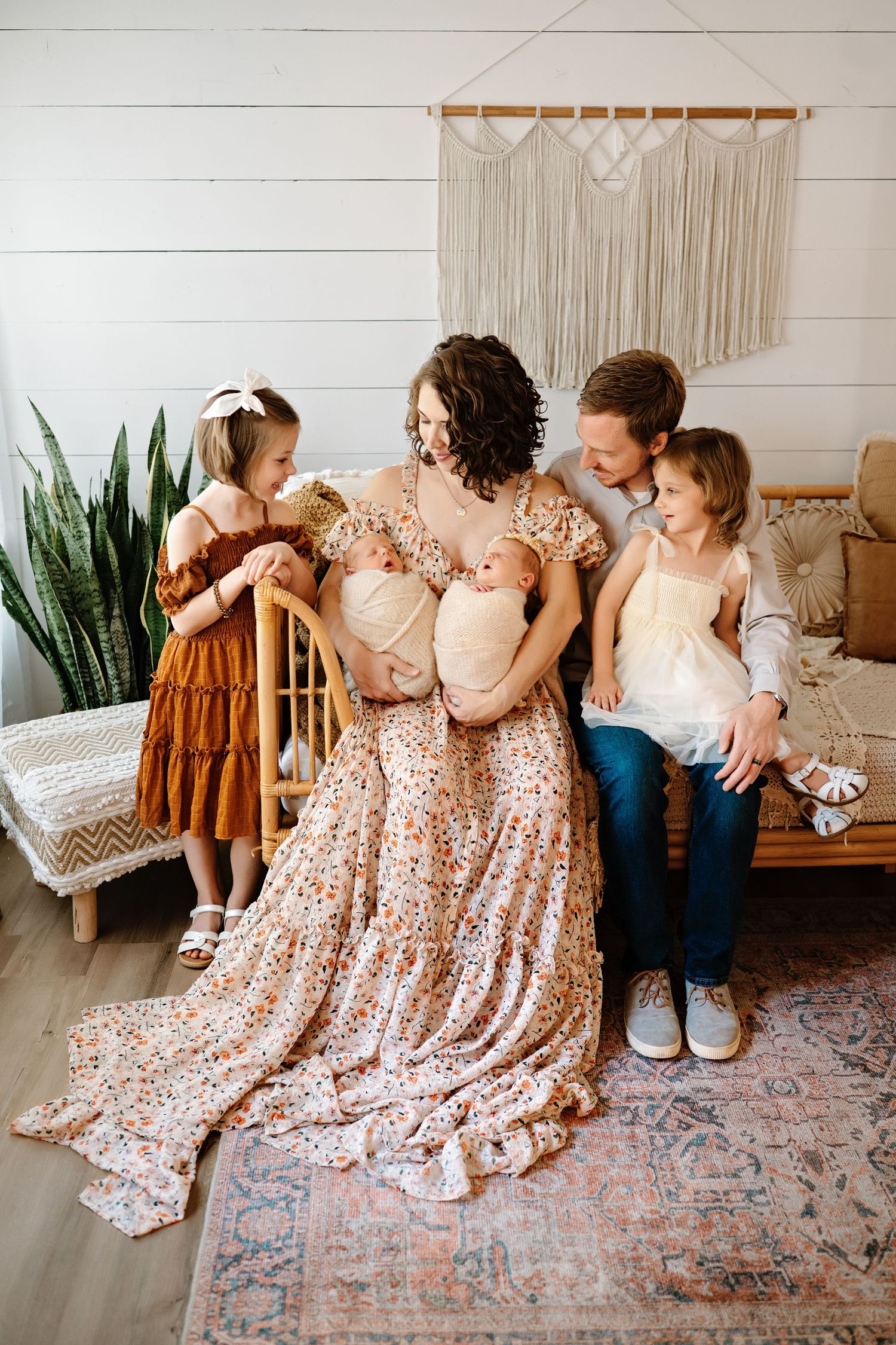 A family sits together on a couch in a cozy boho-style room with macrame wall art and a potted snake plant.