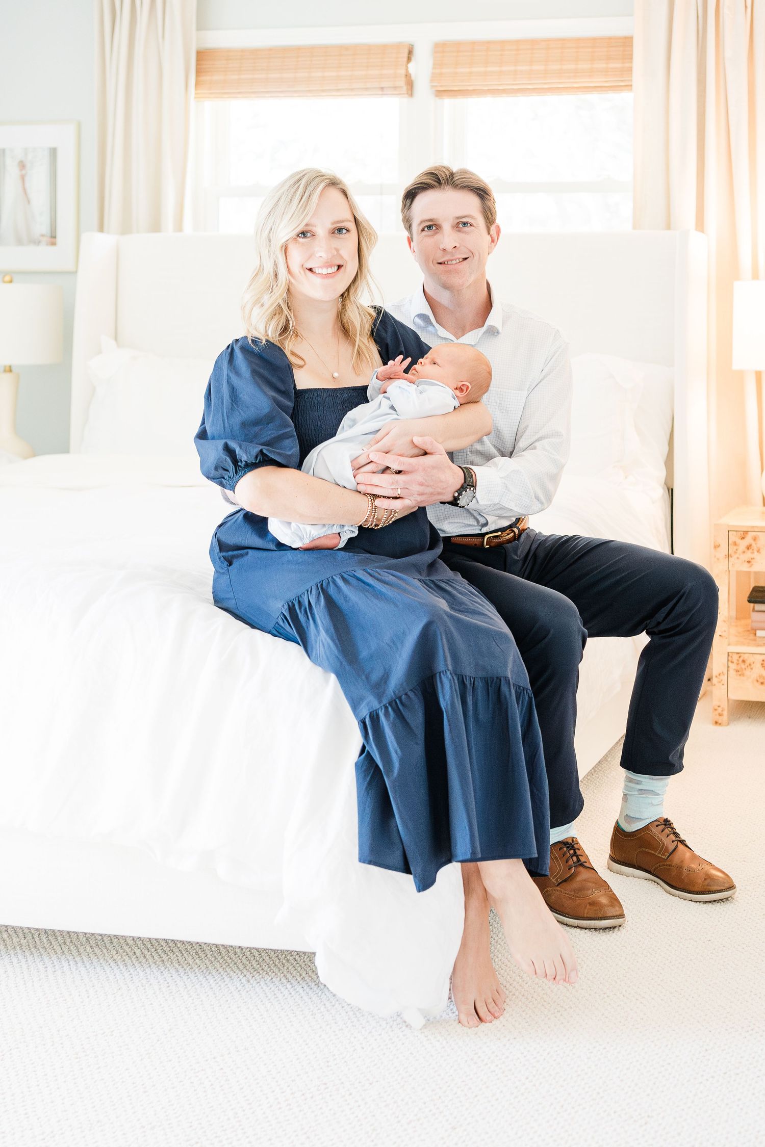 Mom and Dad sitting on their bed holding their baby, looking at the camera and smiling