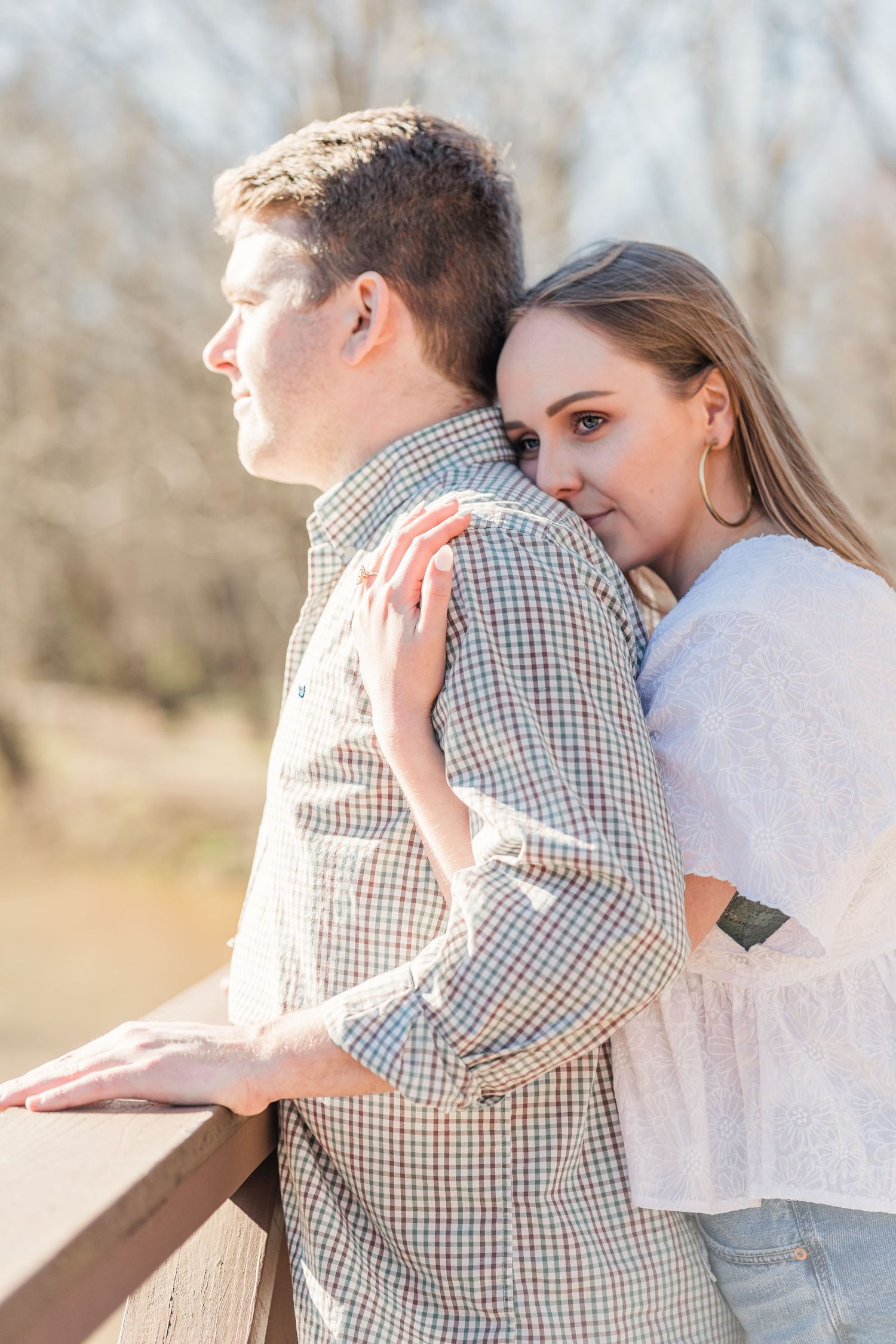 Close up shot of woman standing behind fiancee hugging him looking out over a rail at the river