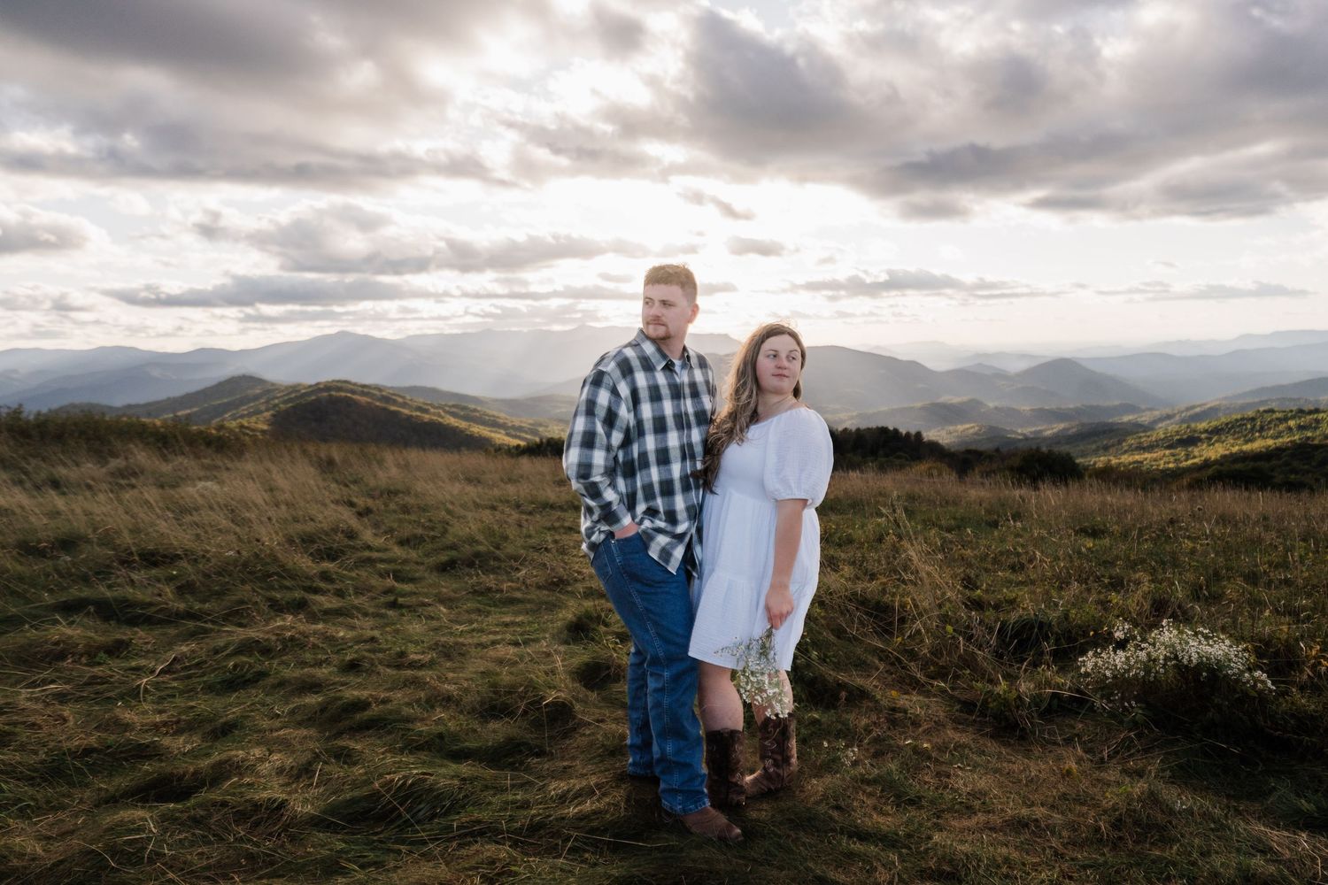 Engaged couple standing hip to hip, facing away from each other. They are wrapped in the golden light of the setting sun on top of Max Patch