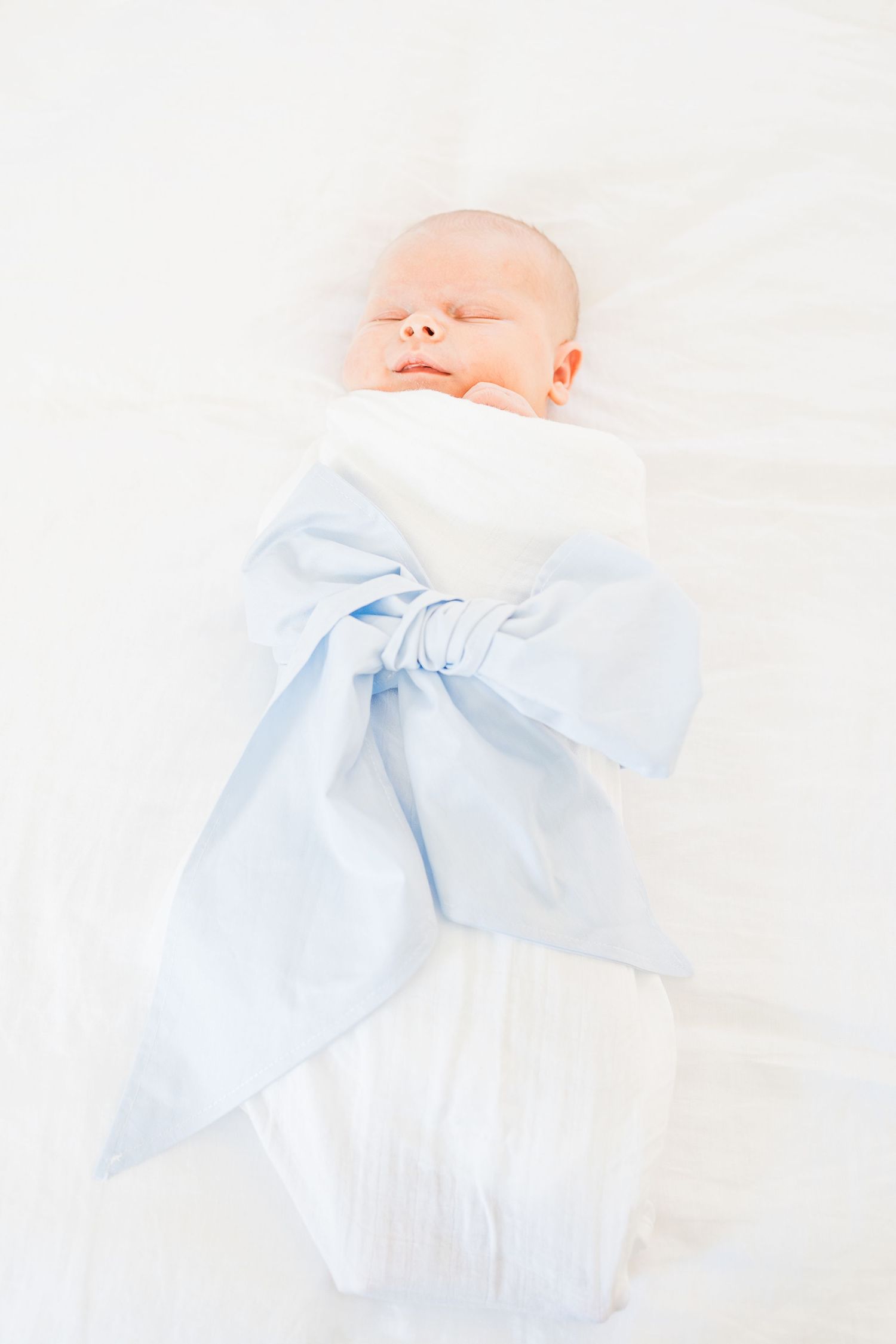 Baby Bradham wrapped in a white swaddle and blue bow lying on his parent's bed during an in-home newborn lifestyle session in Greenville SC