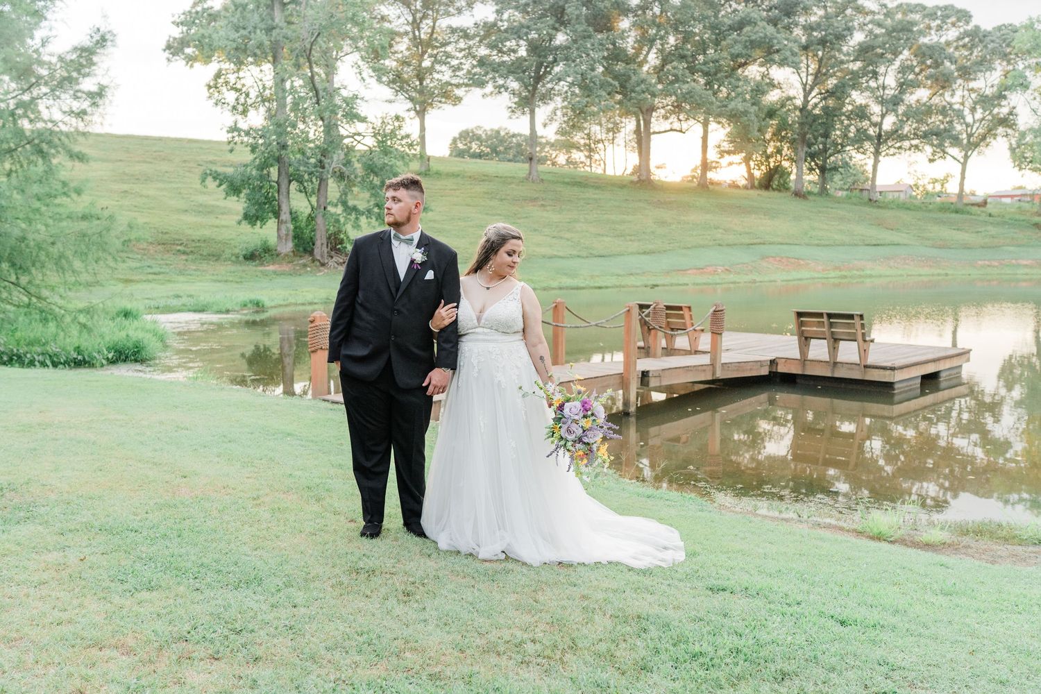 bride and groom standing in front of the lake dock with the golden sun peeking between the trees behind them.