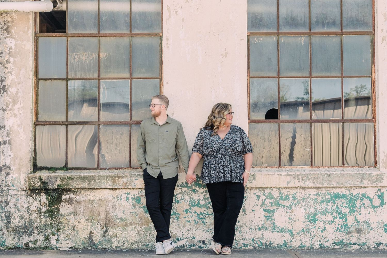 Couple holding hands in front of an industrial wall of windows at Taylors Mill in casual outfits
