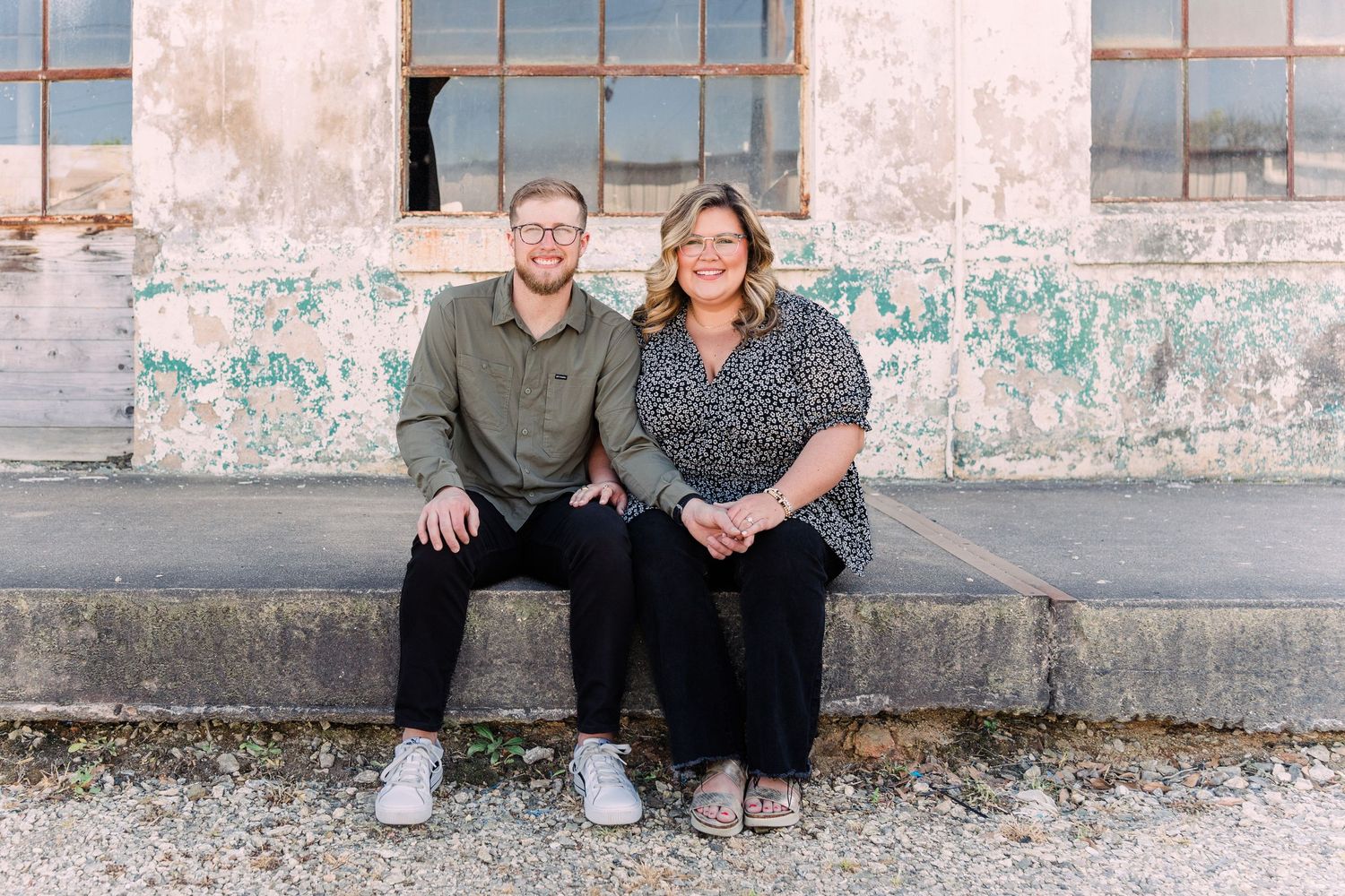 Man and woman sitting down on the sidewalk in front of Taylors Mill industrial windows, holding hands and smiling