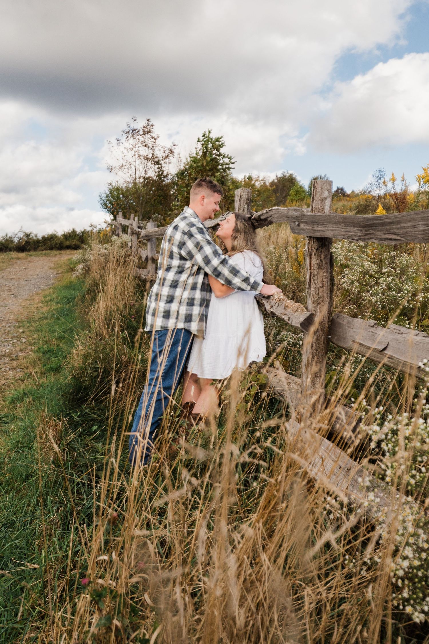 Engaged couple leaning against a rustic wooden fence staring lovingly into each other's eyes.