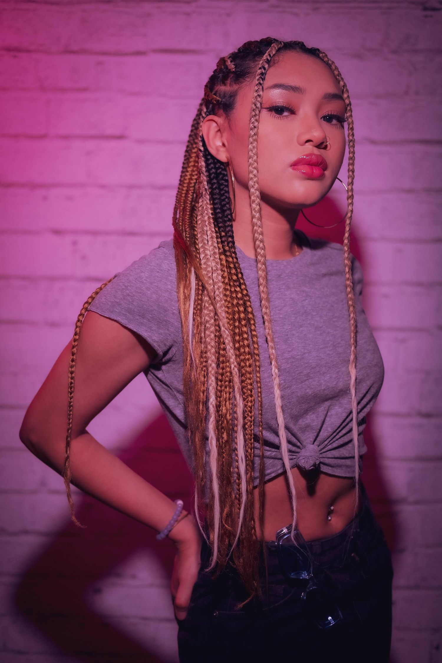 Model with long braids posing stylishly against purple-lit brick wall background.