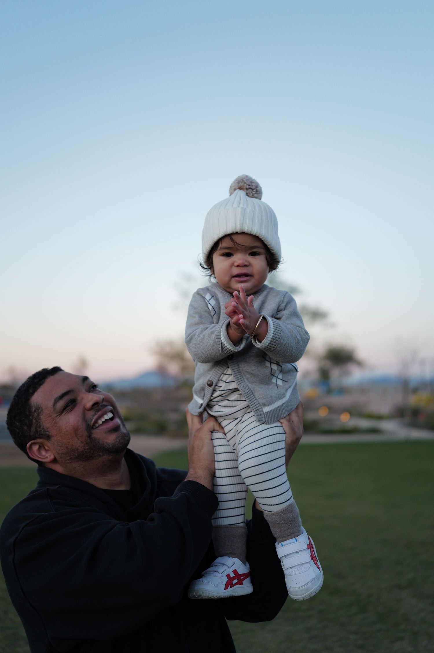 Parent lifting small child wearing winter hat during sunset in a park.