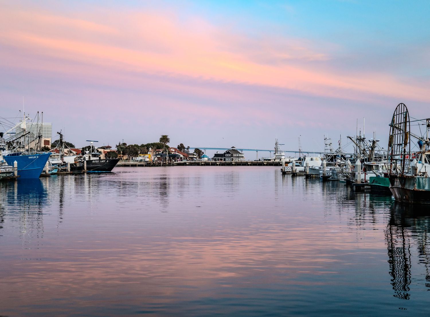 Marina at sunset with boats and fishing vessels docked along the waterfront under pink skies.