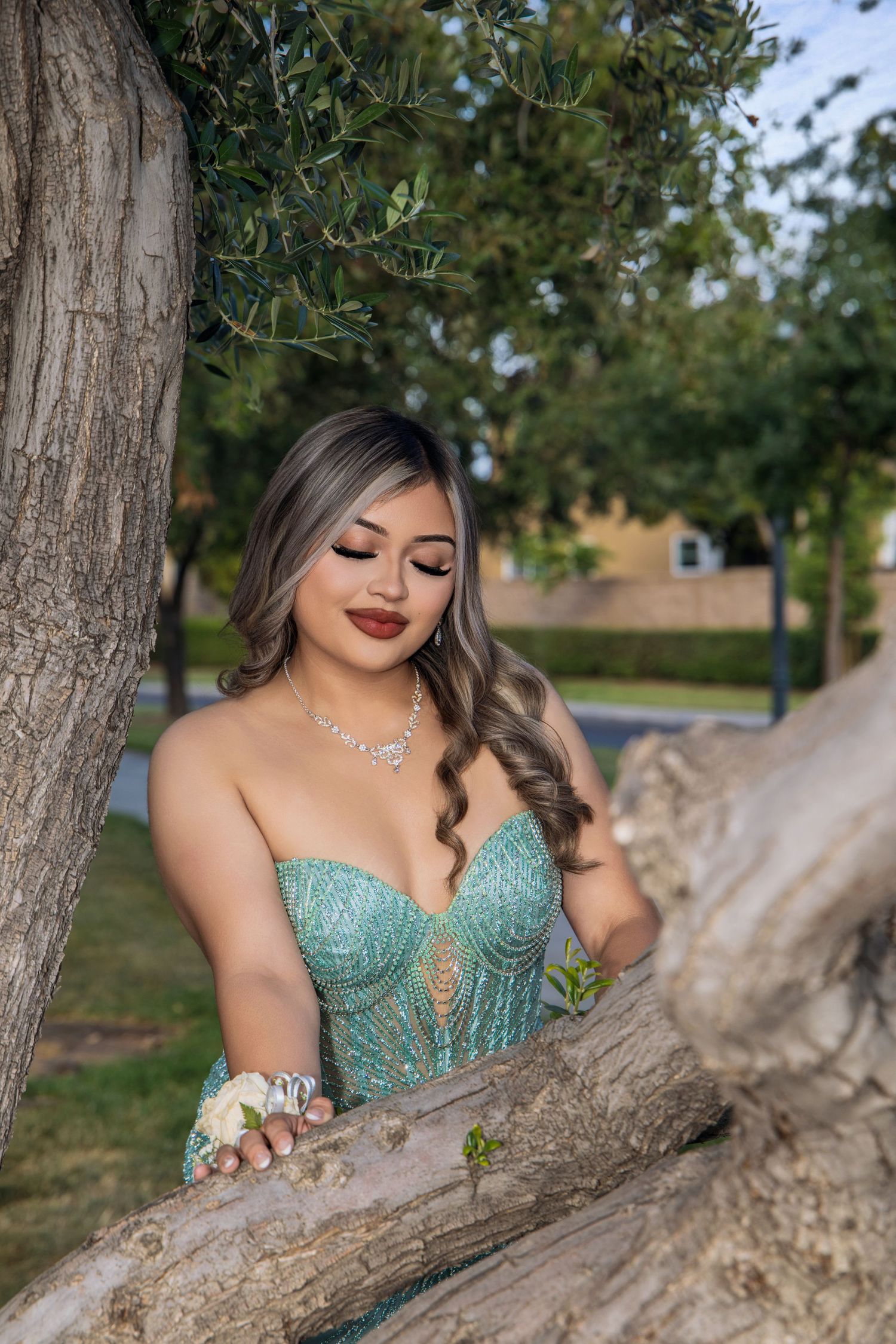 Person in teal formal dress posing elegantly by tree trunk in outdoor setting.