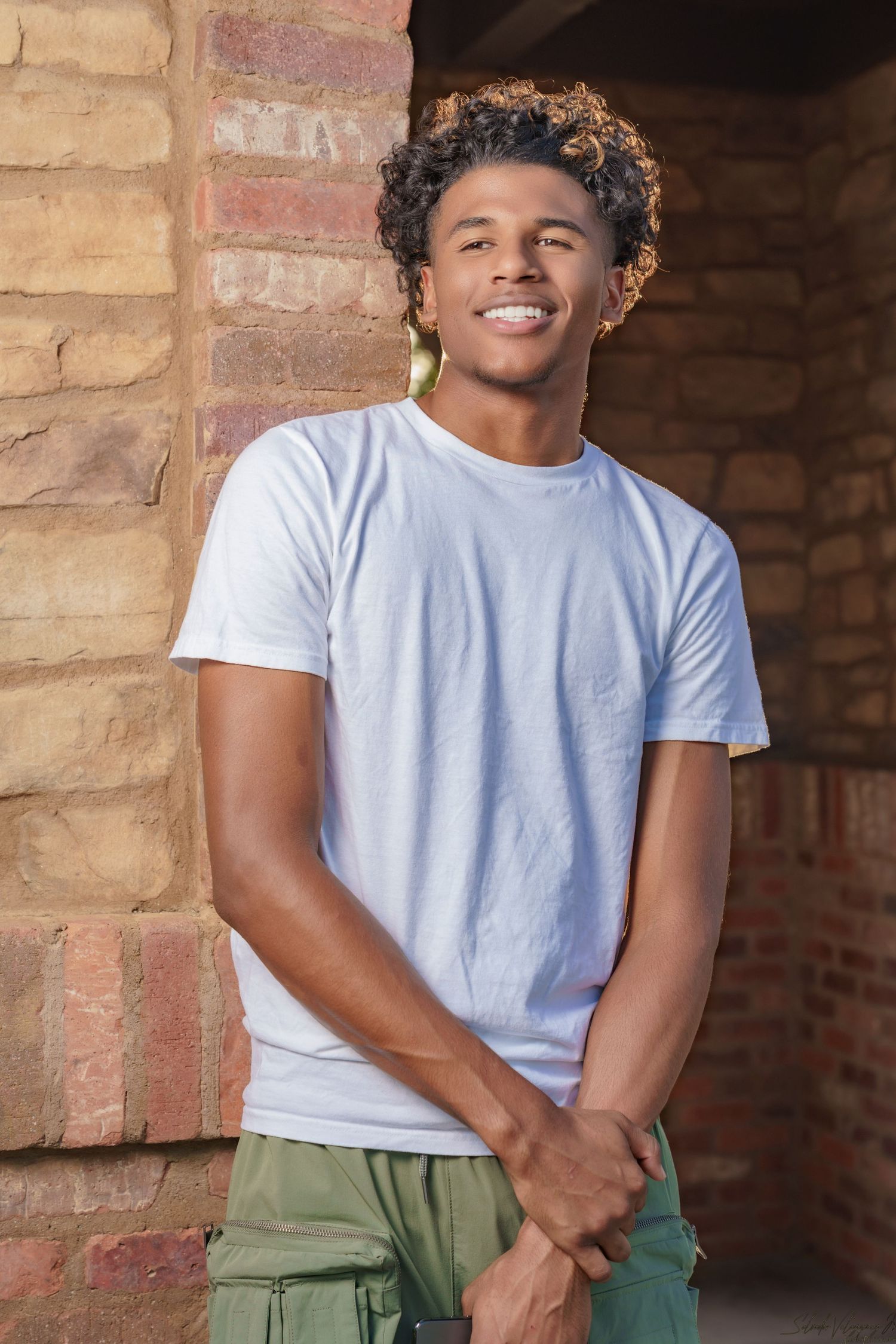 Person in white t-shirt and green pants posing casually against brick wall.