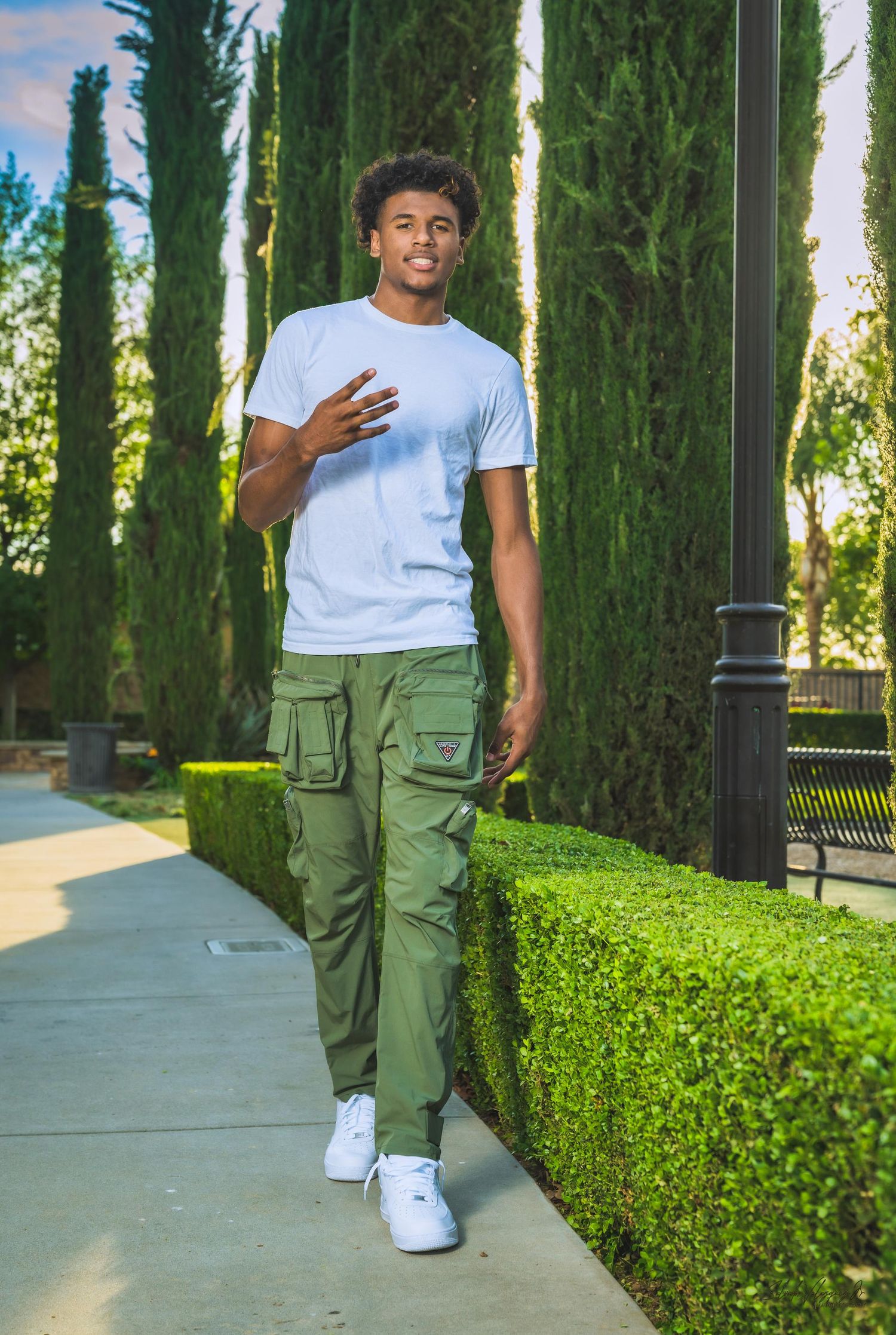Young person in white t-shirt and cargo pants standing between tall cypress trees.
