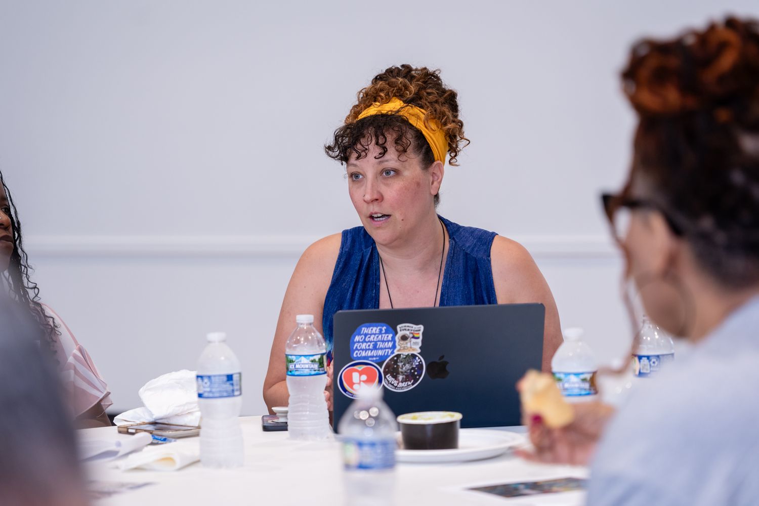 Woman in blue dress speaking at conference table with laptop covered in stickers.