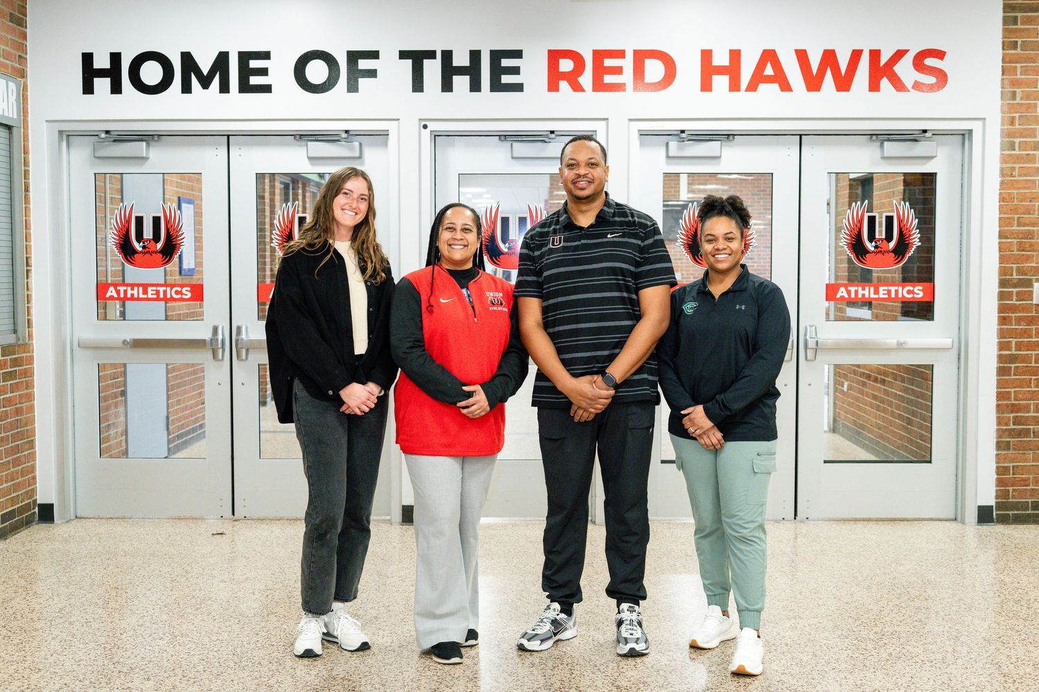 Four people posing together in front of a Home of the Red Hawks school athletics entrance.