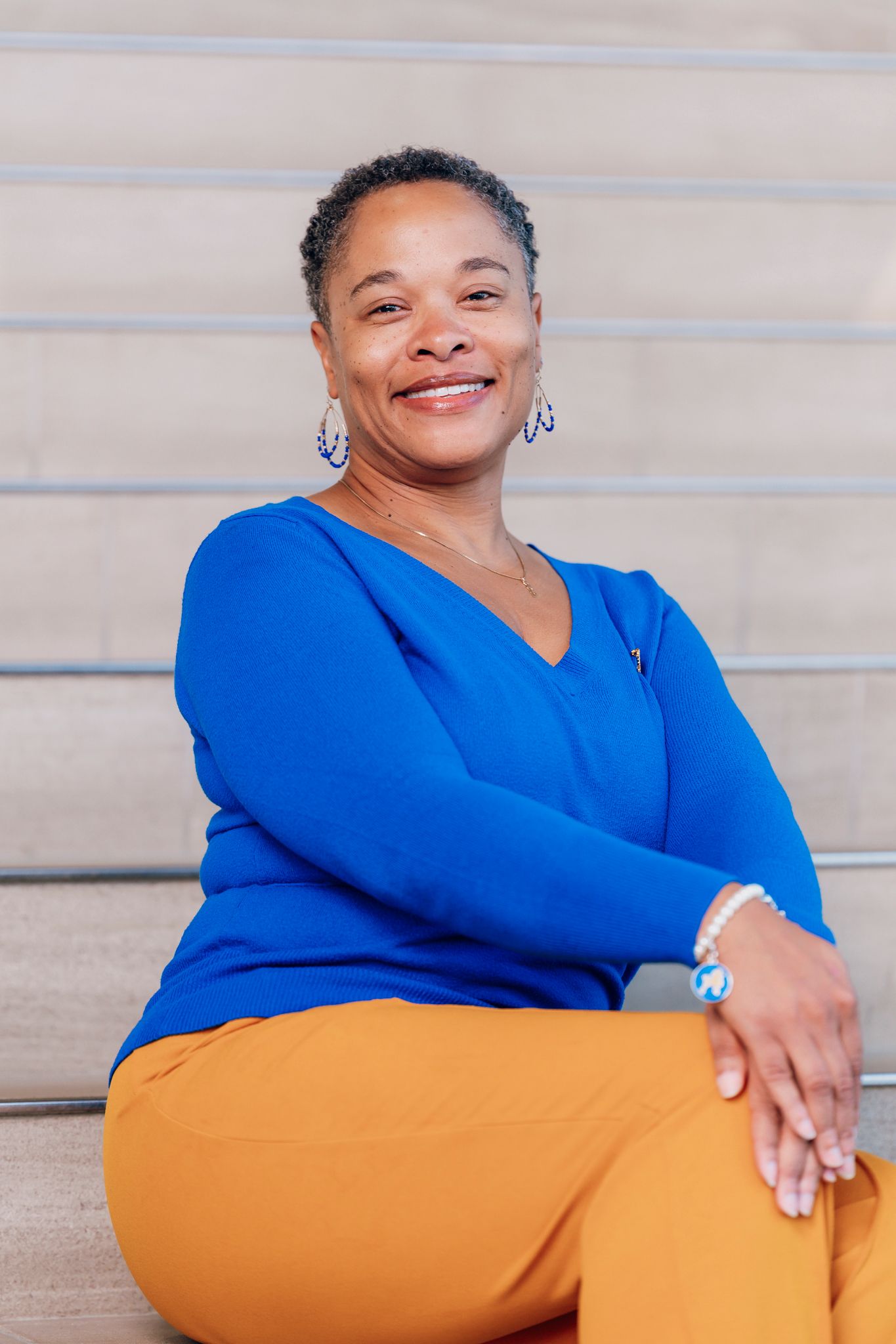 Smiling woman in a royal blue top and mustard pants sits on light stone steps, wearing drop earrings, a bracelet and a necklace.