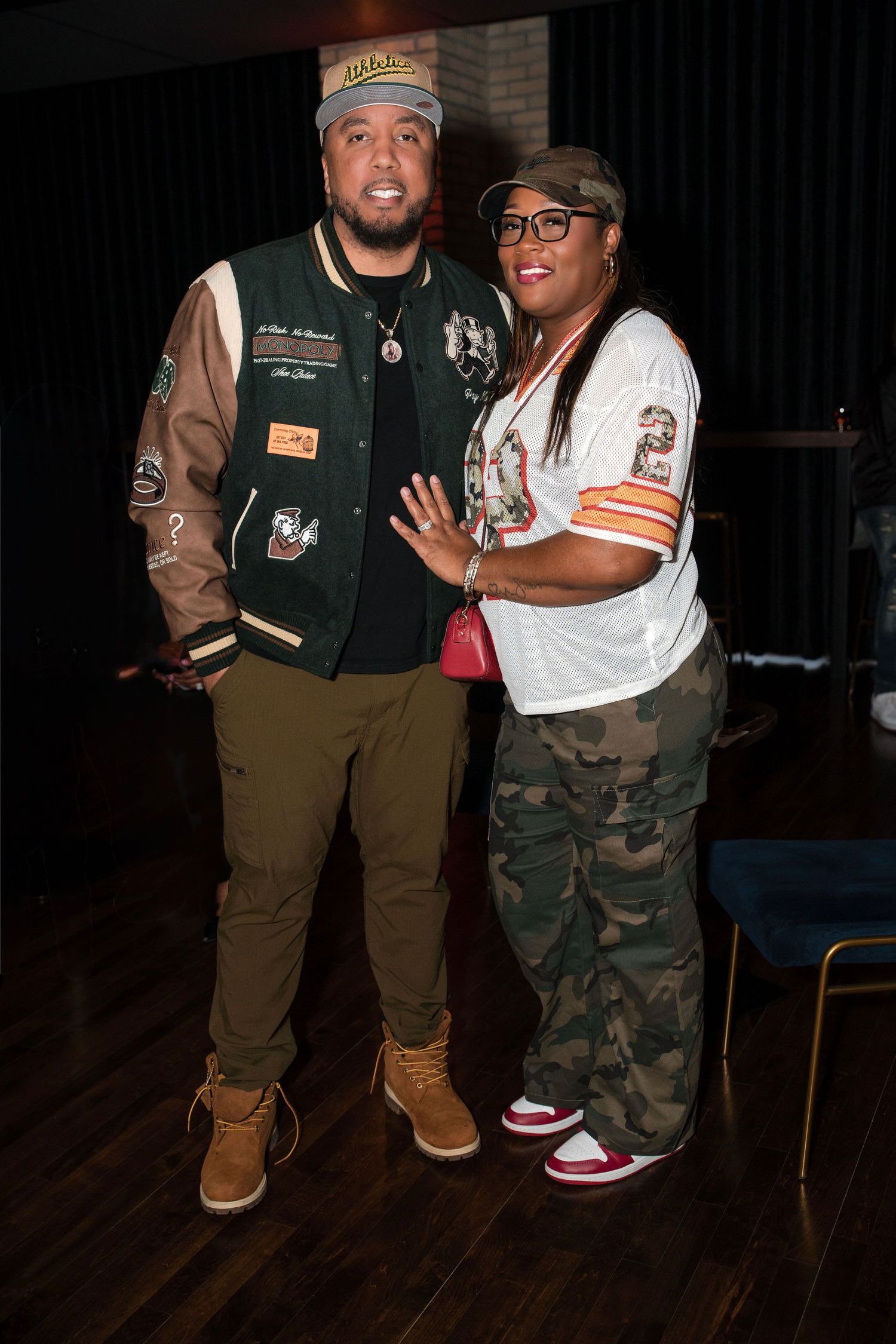 Two people posing together indoors, wearing casual streetwear including varsity jacket, camo pants, and sneakers.