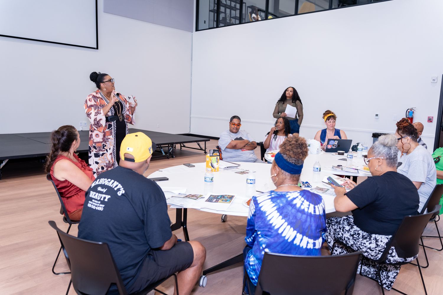Diverse group of people seated at a round table during a workshop, with a presenter standing and speaking.
