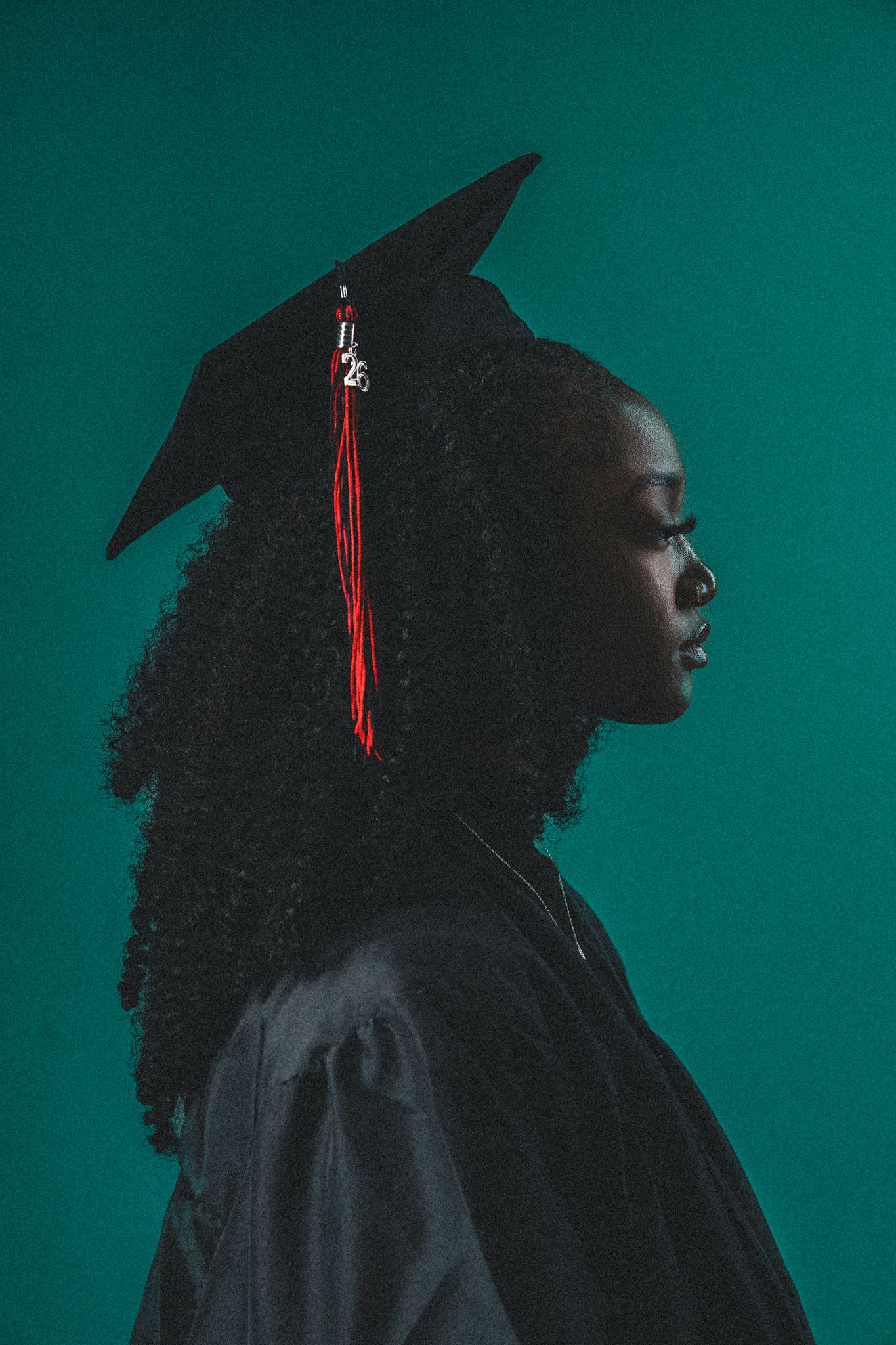 A young Black woman in a black graduation cap and gown poses in profile against a teal background.