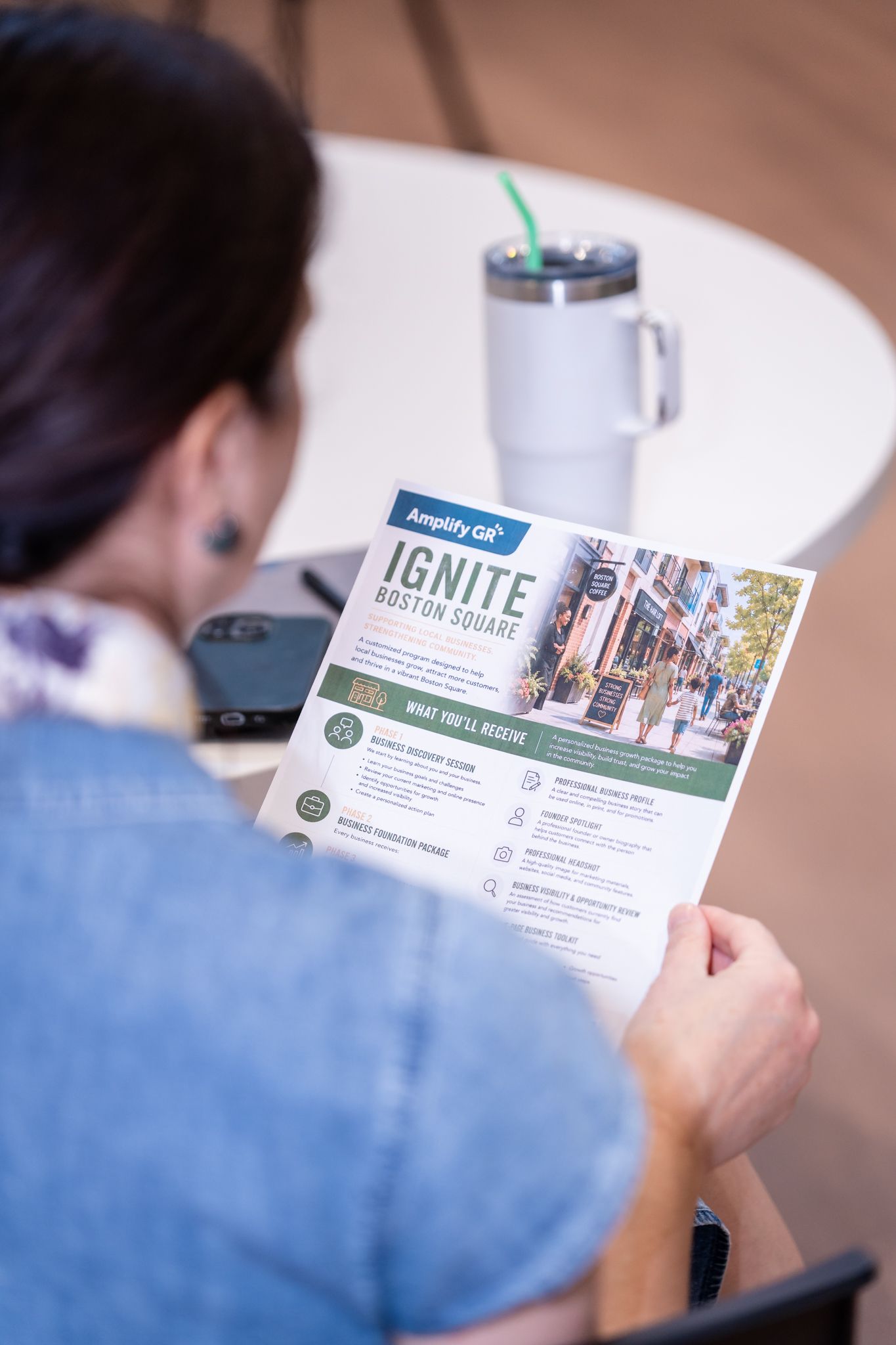 Woman reading an Ignite Boston Square flyer from Amplify GR at an outdoor cafe table with a tumbler cup.