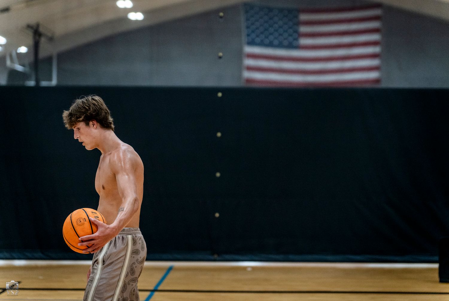 Shirtless young man dribbling a basketball on an indoor court with an American flag hanging in the background.