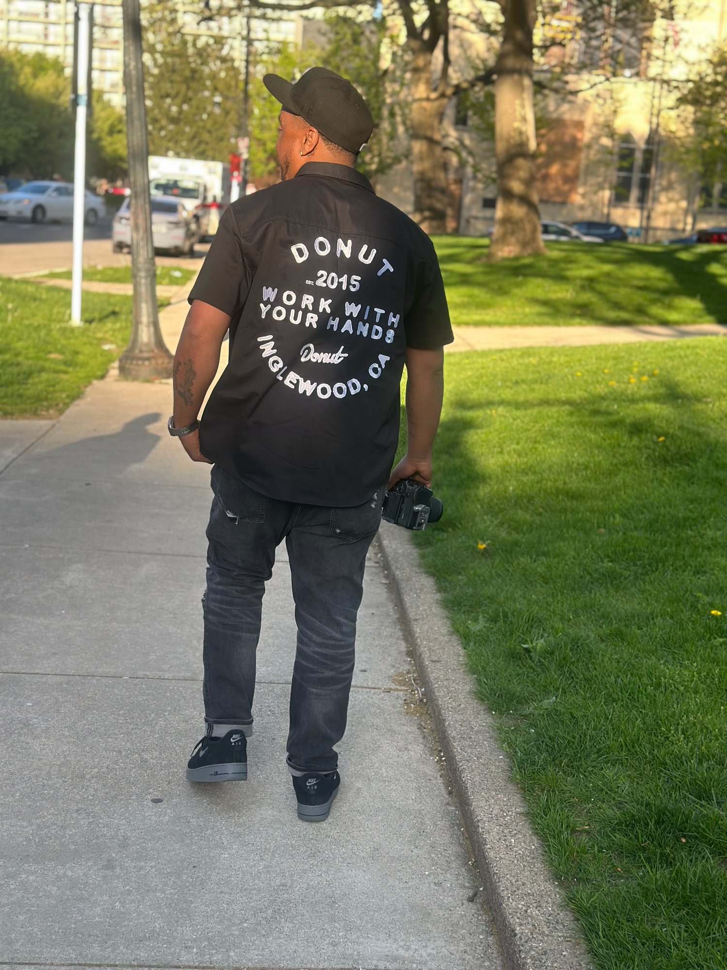 Man walking on sidewalk wearing black Donut 2015 Work With Your Hands Inglewood CA t-shirt and hat.