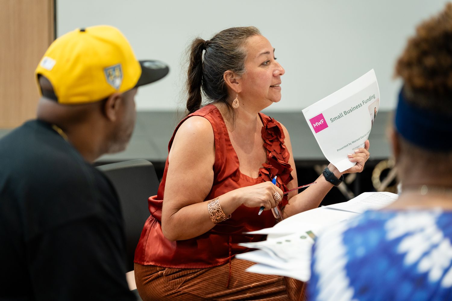 Woman in red dress holds papers, speaking at a meeting with diverse attendees in a bright conference room.