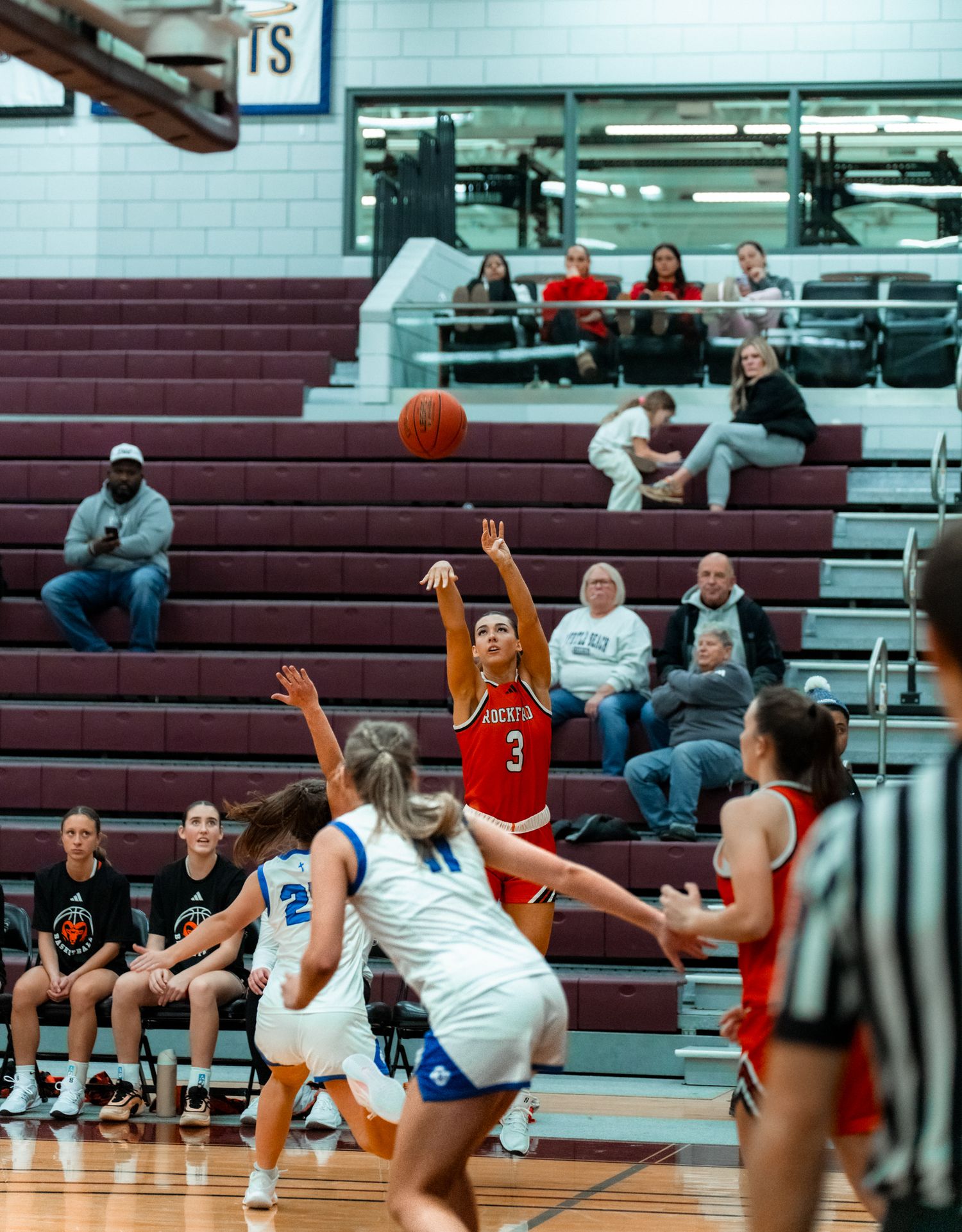 Girls basketball game with player #3 in red shooting over defenders in blue and white uniforms in a school gymnasium.