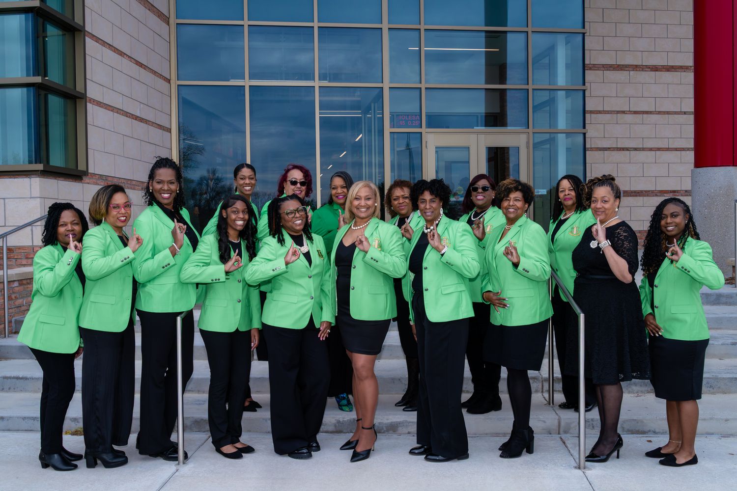 Group of women in matching green jackets and black skirts or pants posing together on the steps outside a modern glass-front building.