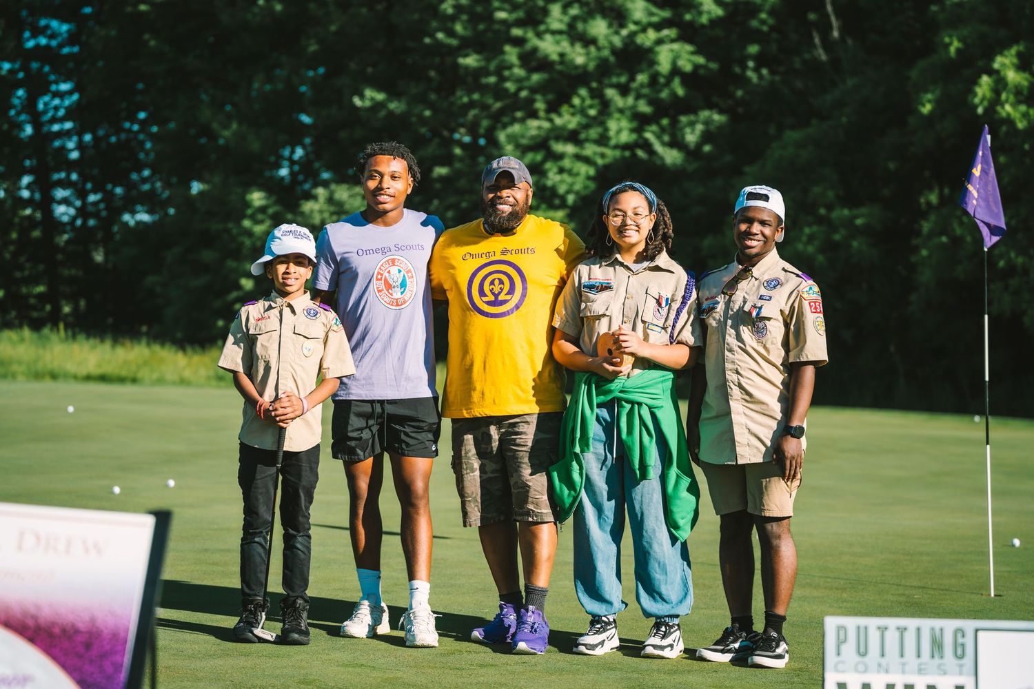 Five smiling people in scout-style uniforms and casual shirts pose together on a green golf course near a purple flag and putting sign.