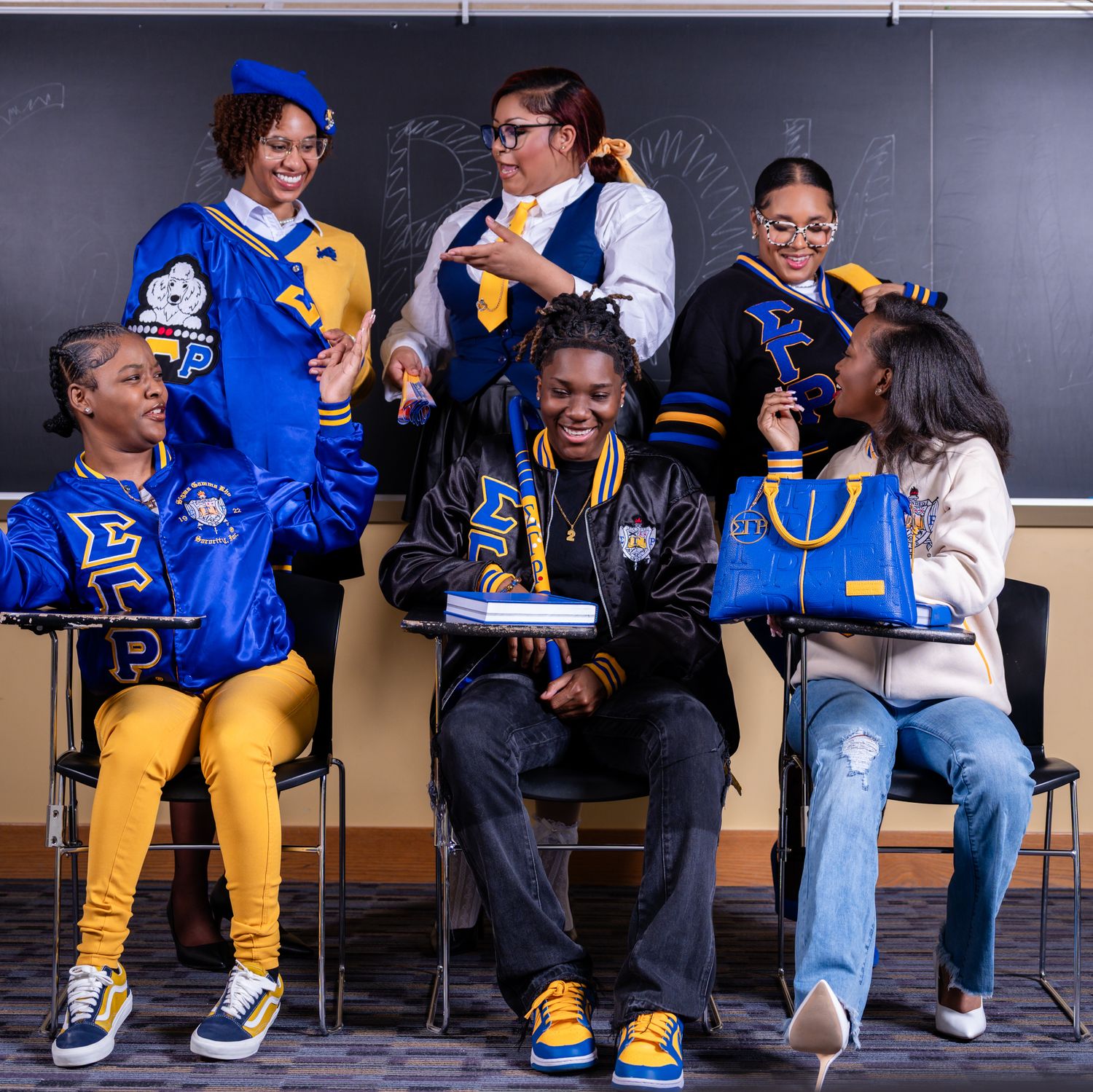 Five students in blue and gold varsity jackets and Greek letter apparel pose together in a classroom with a chalkboard behind them.