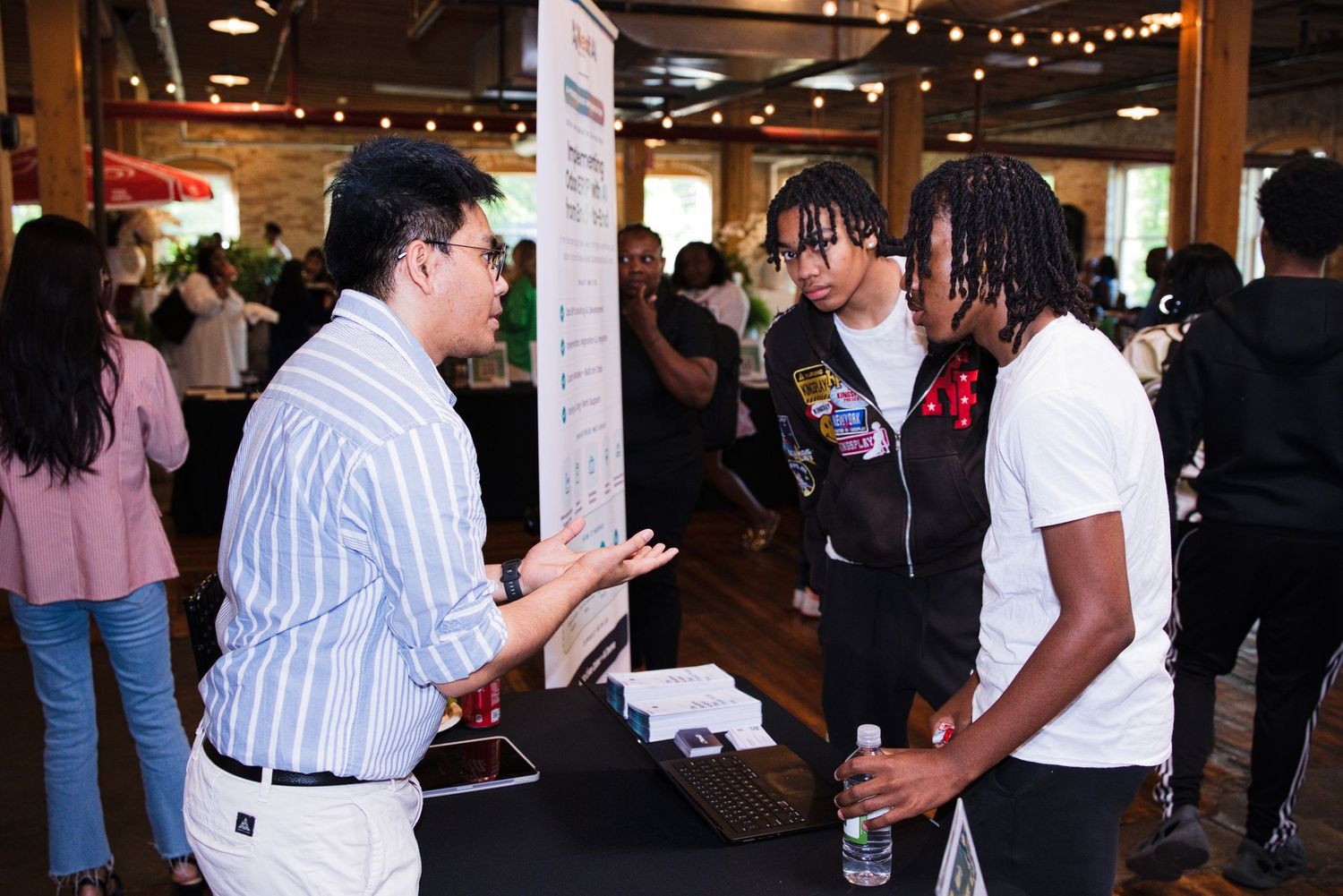 A man in a striped shirt speaks with two young men at an informational booth during a community event.