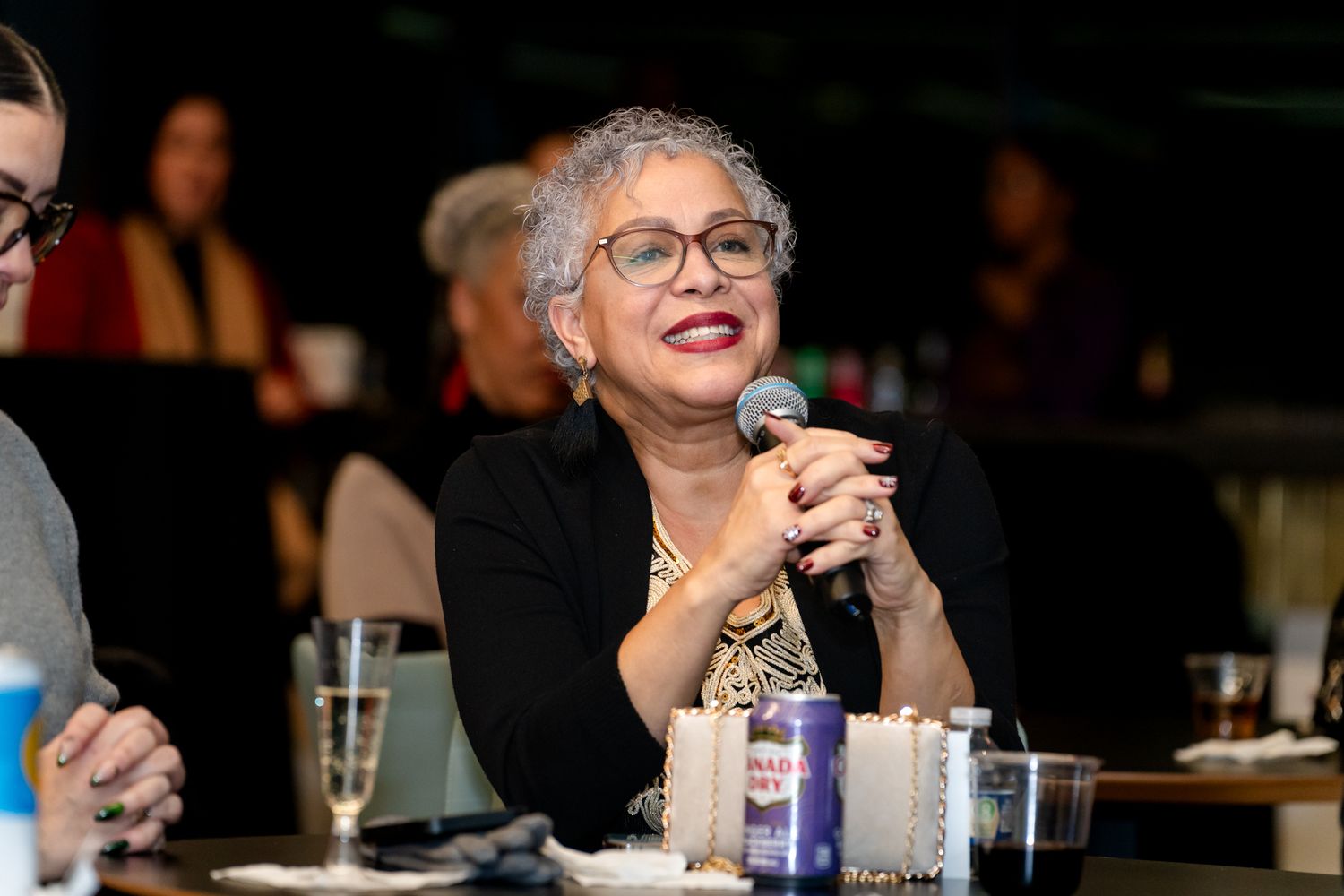 Older woman smiling and speaking into a microphone at a formal dinner event.