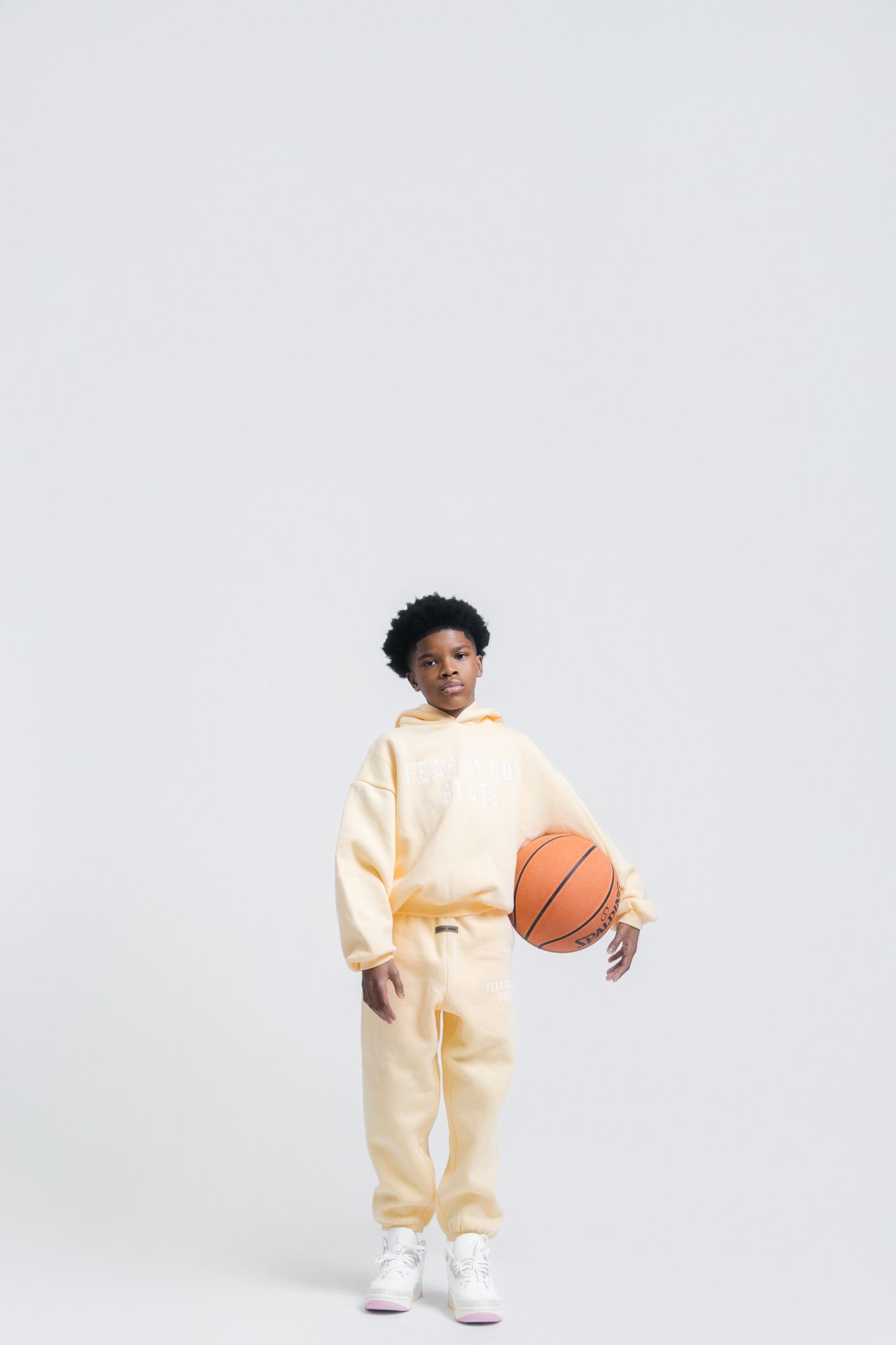Black boy in yellow hoodie and sweatpants holds basketball on white background.