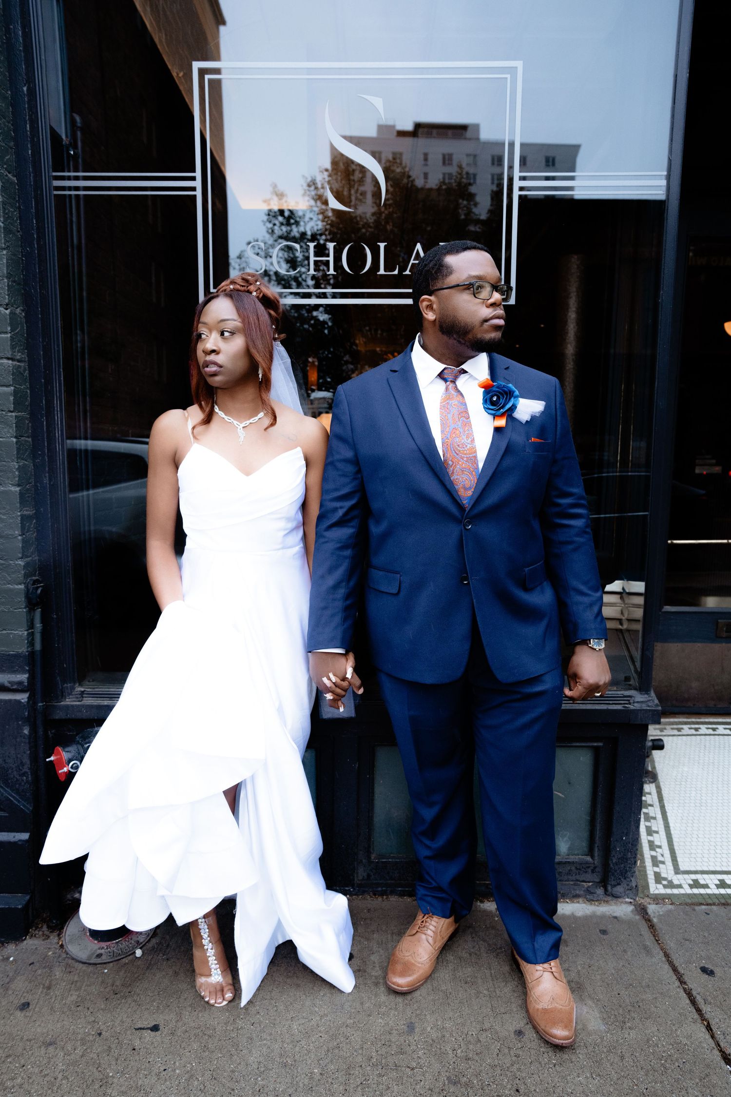 Bride and groom in wedding attire posing outside Schola restaurant on urban sidewalk.