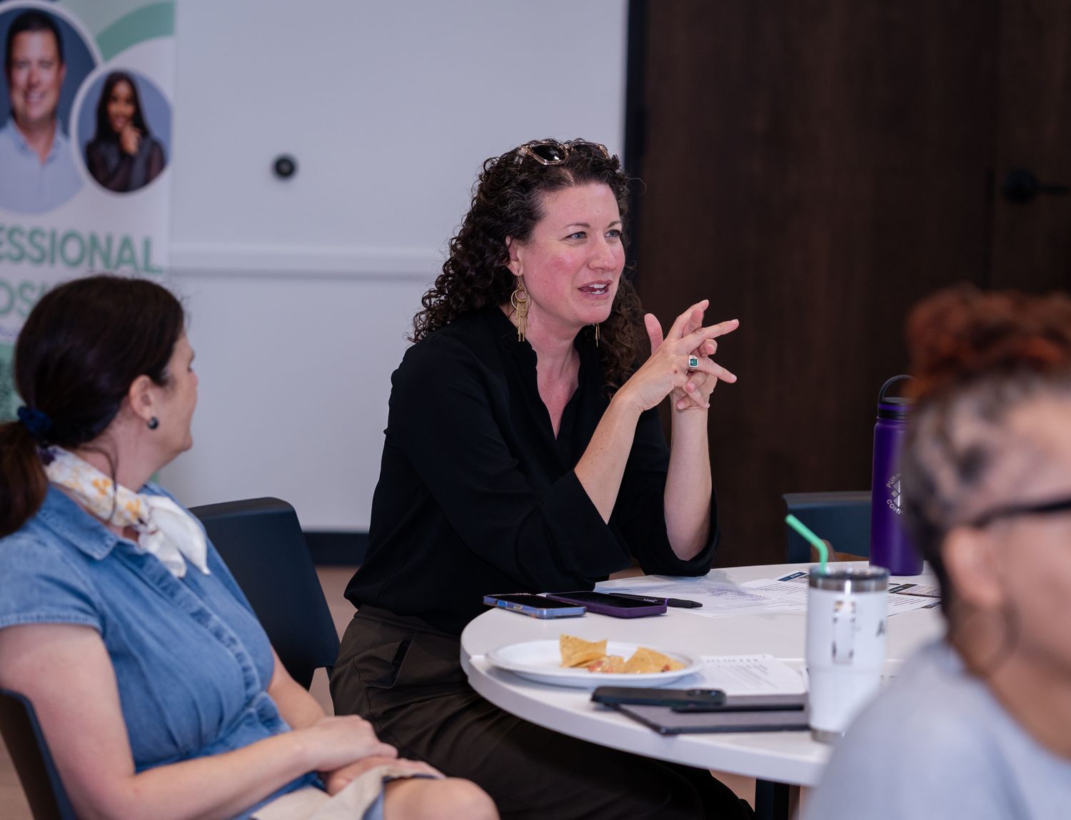 Professional woman speaking and gesturing at a business networking event with attendees seated at a round table.