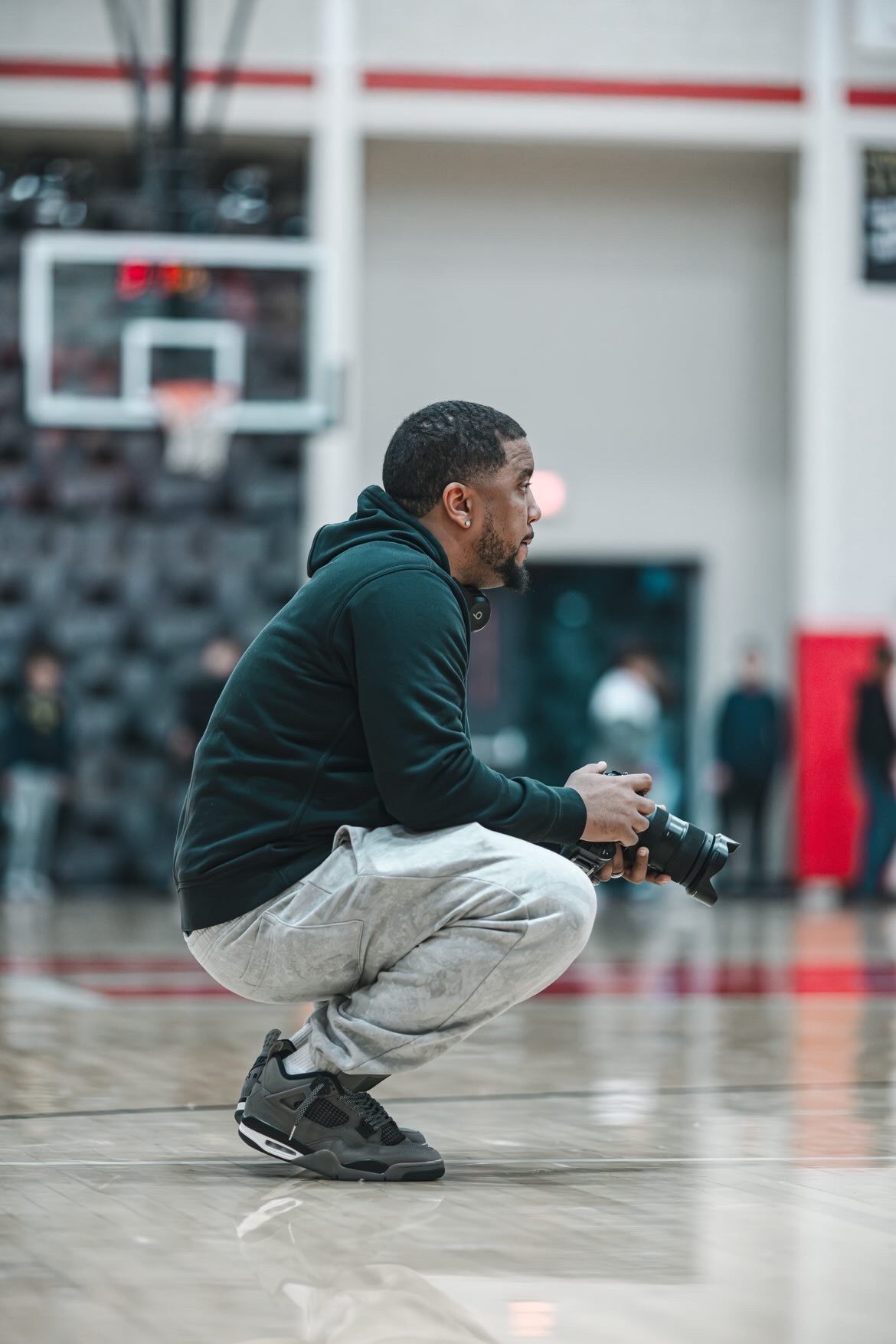 A photographer crouches on a basketball court holding a camera, wearing a dark hoodie, gray sweatpants, and black Jordan 4s.