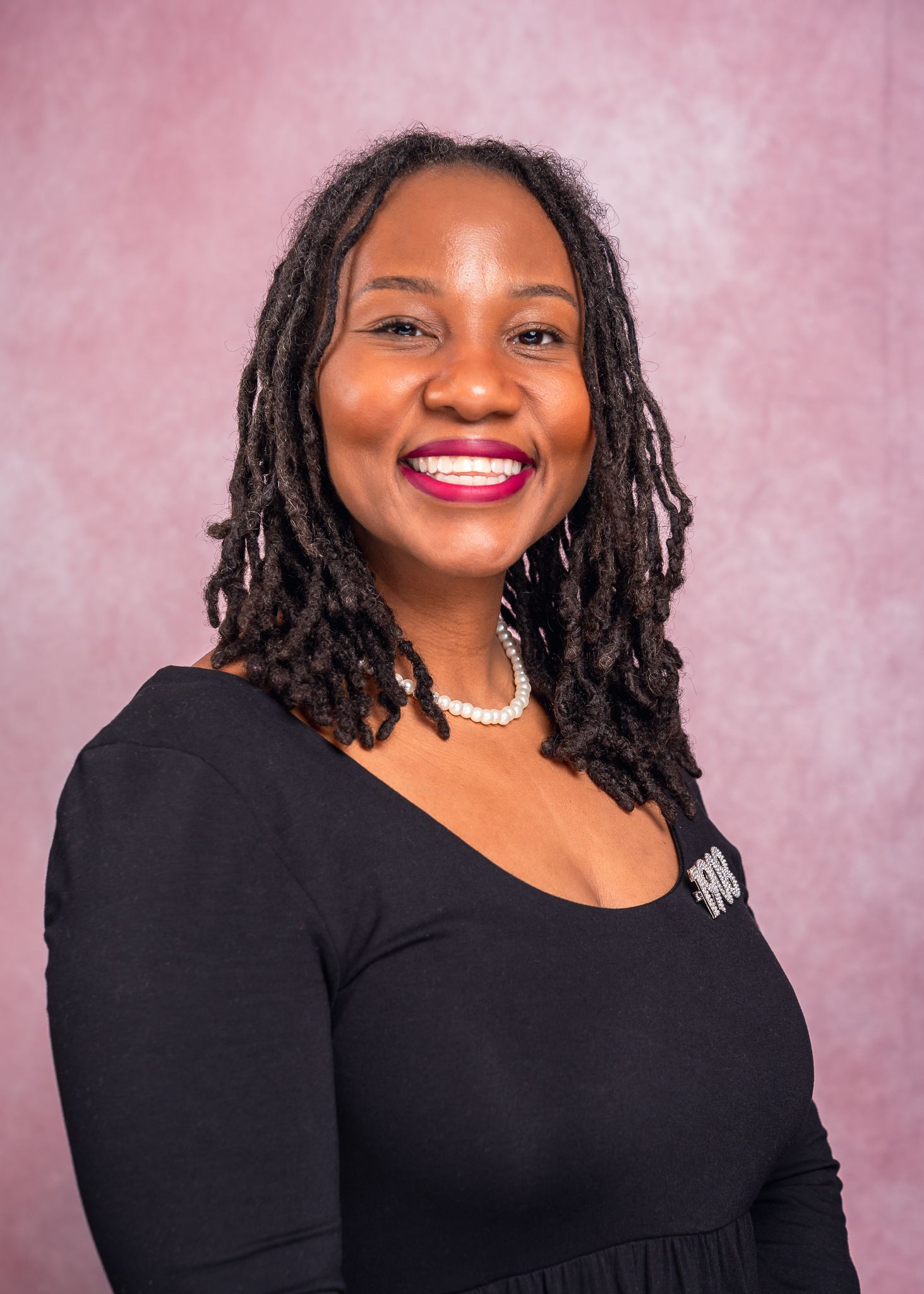 Smiling woman with shoulder-length locs wears a black top and pearl necklace in a professional portrait against a pink backdrop.