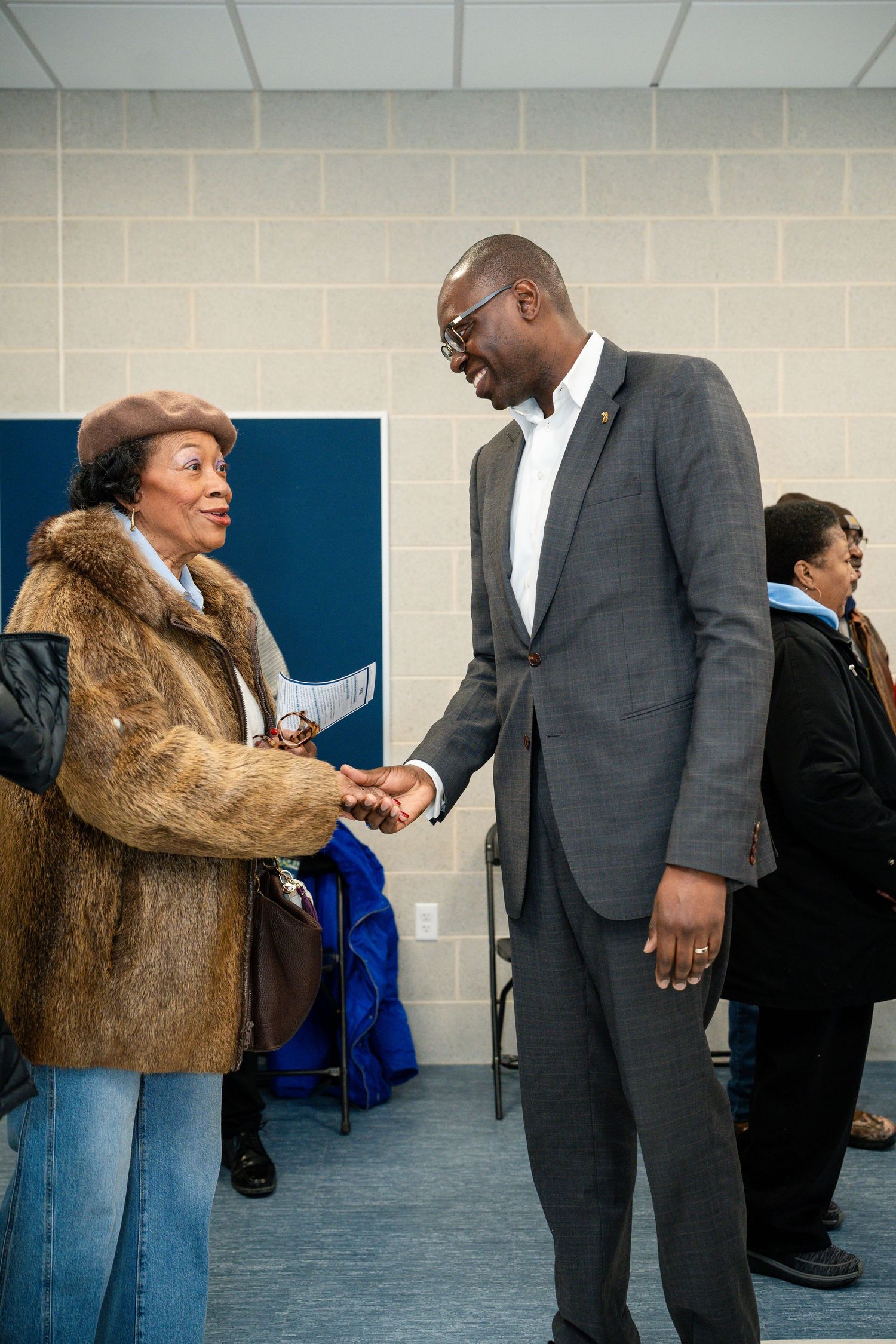 A tall man in a gray suit shakes hands with an elderly woman in a fur coat at a community gathering.