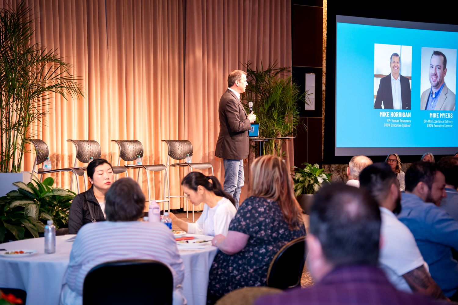 Speaker presenting to seated attendees at the Amway GROW business event with panelist photos on screen.
