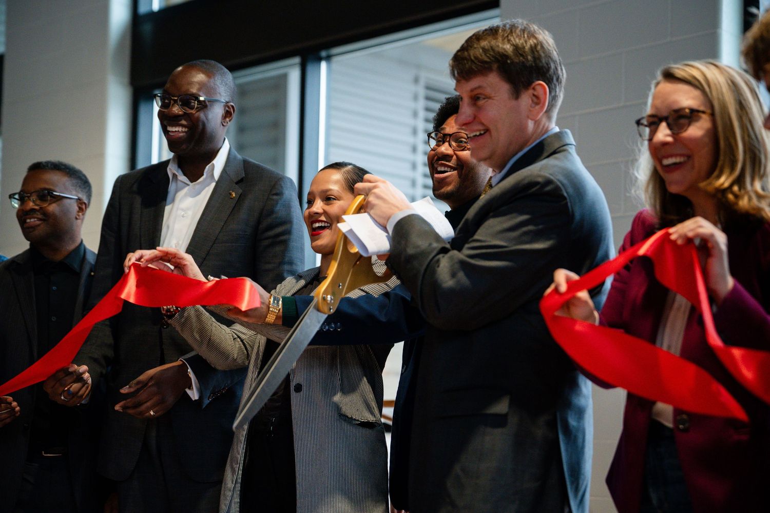A diverse group of smiling professionals cuts a red ribbon at a formal grand opening ceremony indoors.